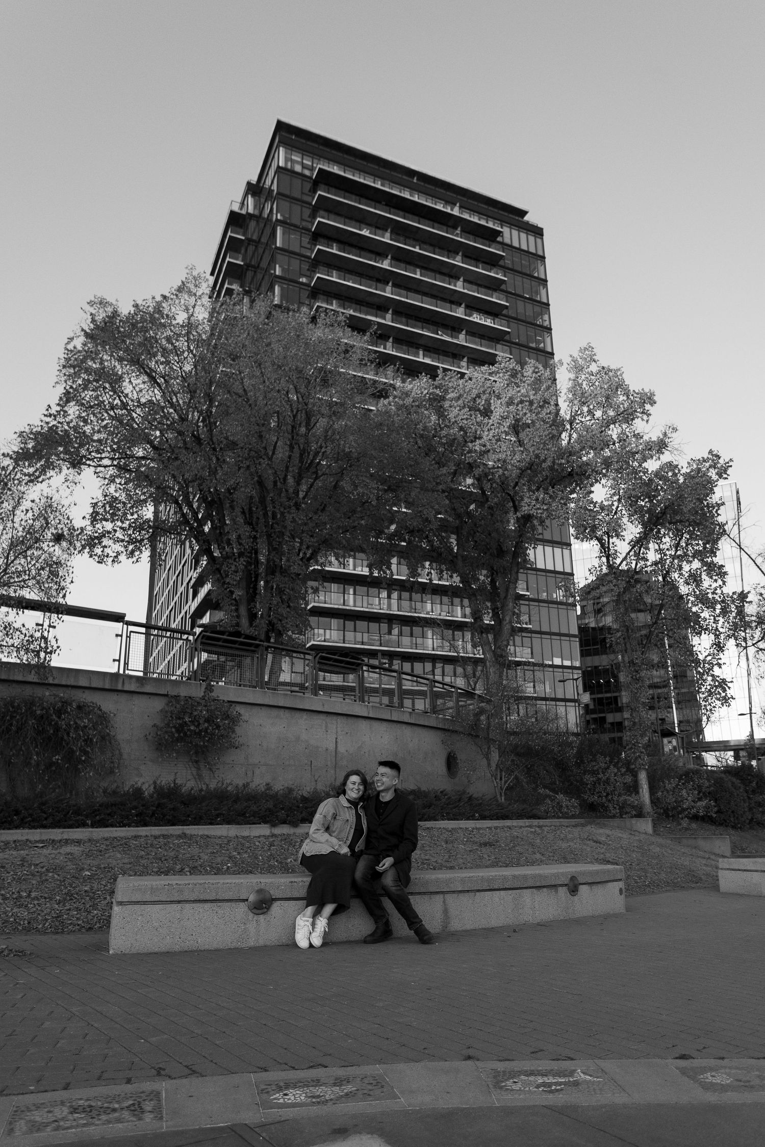 Black and white photo of a couple sitting on a bench with a modern high-rise building and trees in the background.