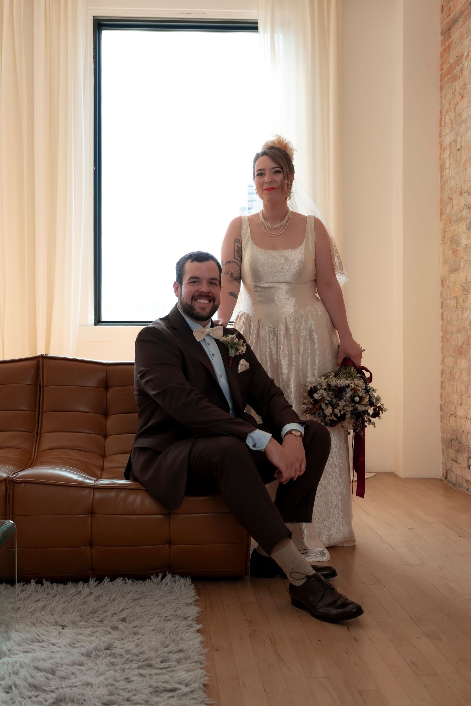 A smiling groom in a brown suit sits on a leather sofa beside his tattooed bride in a white dress holding a bouquet.