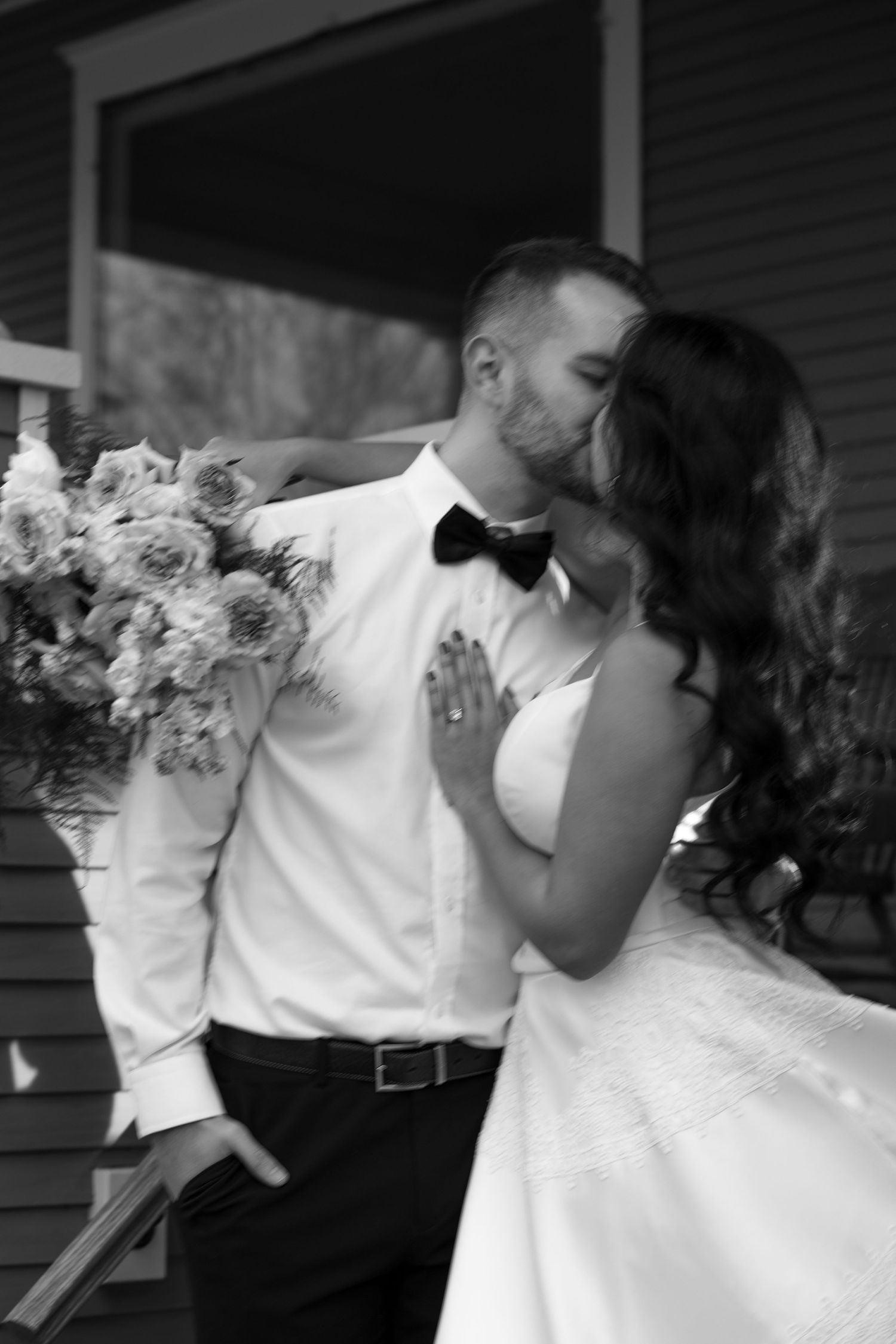 Black and white photo of a bride and groom sharing a kiss, bride holding a floral bouquet, wearing wedding attire.