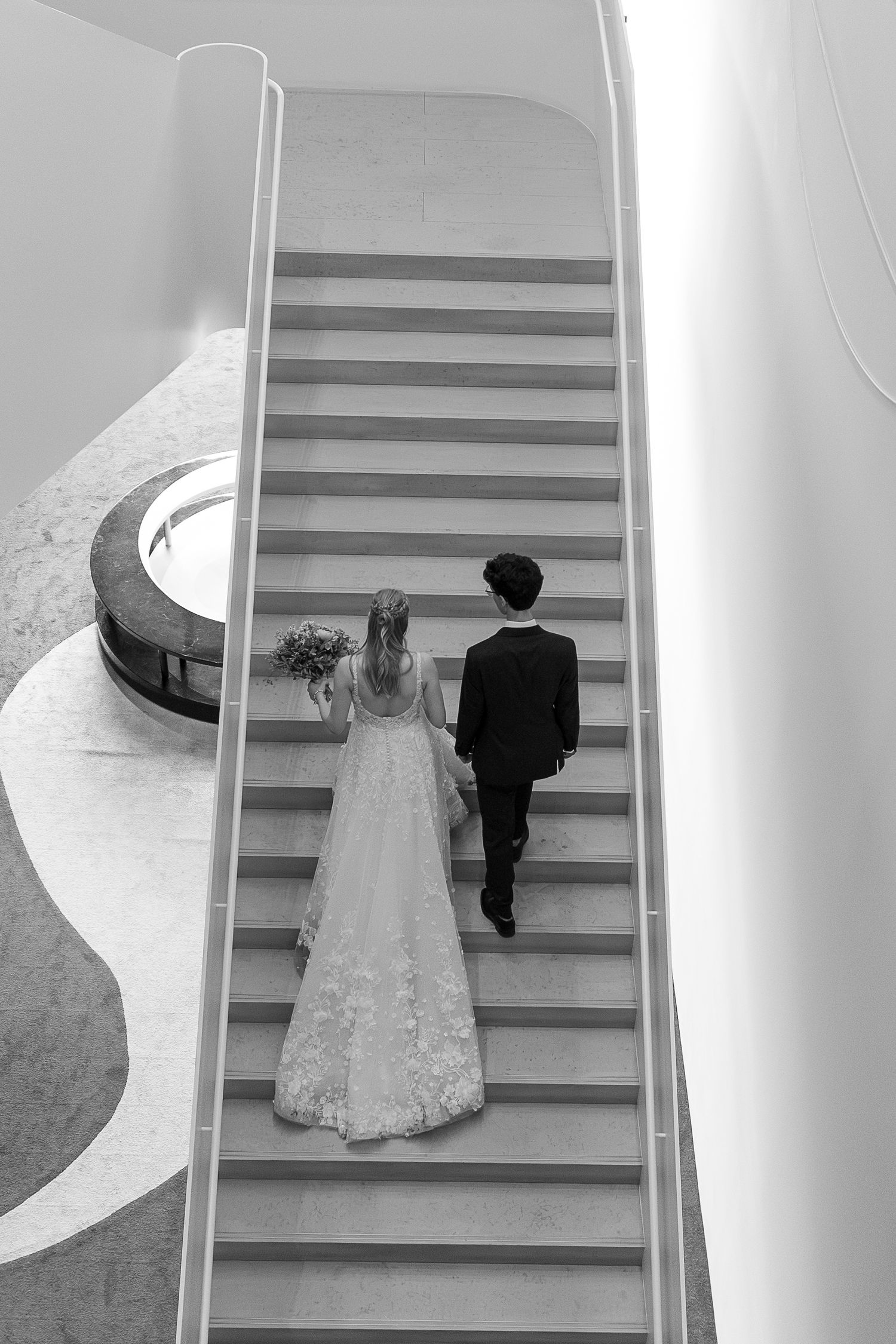 A couple descends elegant white stairs in formal wedding attire at the Remai Art Gallery in Saskatoon, Saskatchewan.