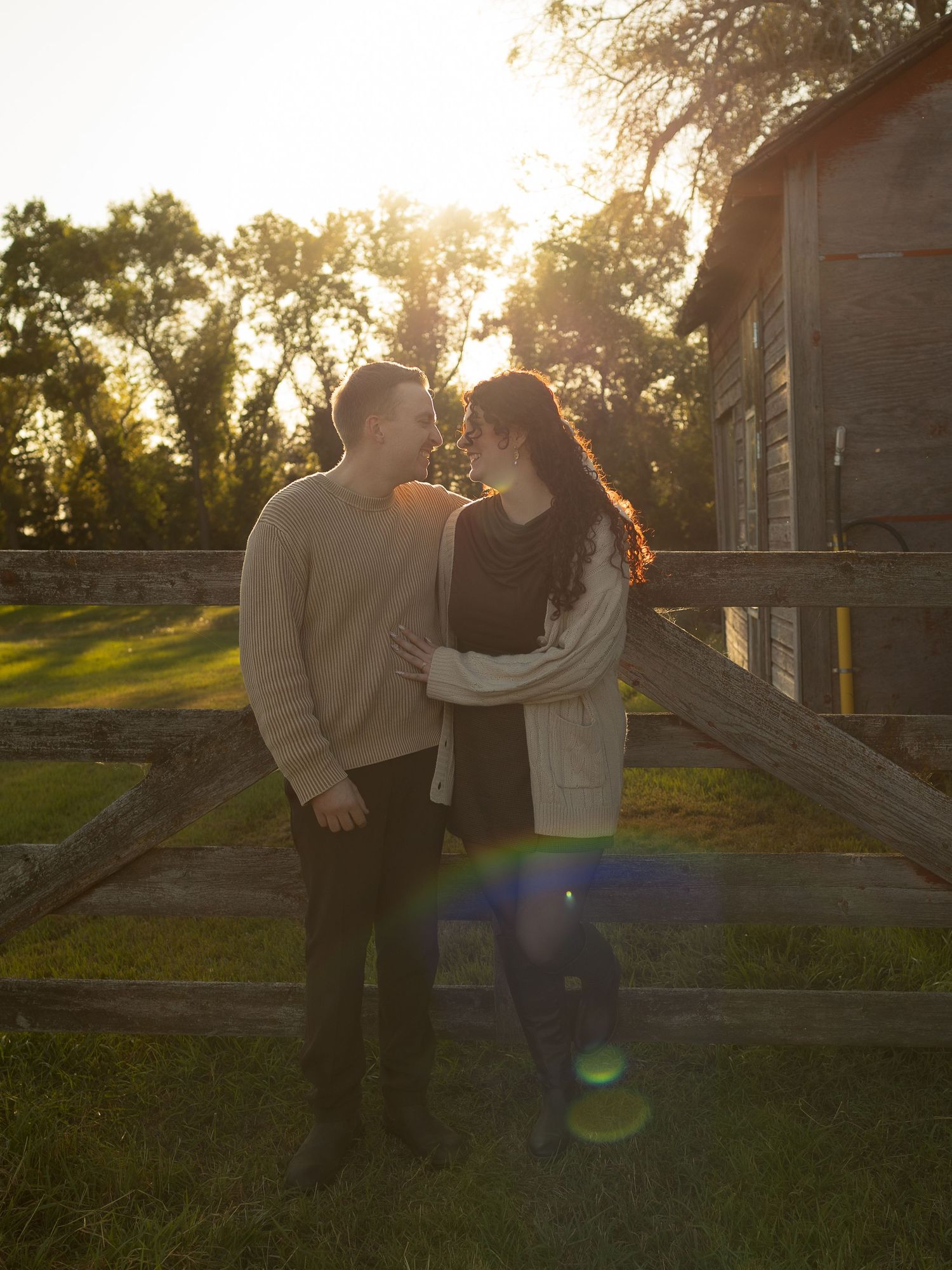 A romantic couple embraces in front of a rustic barn at golden hour with beautiful sunlight streaming through trees.