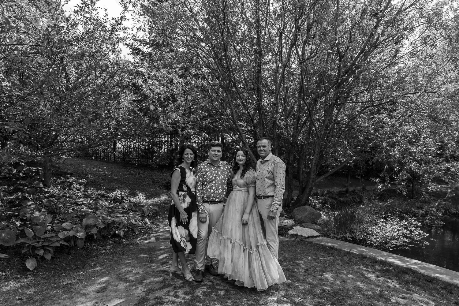 Black and white wedding photo of a bride in a gown with family members posing outdoors among lush trees.
