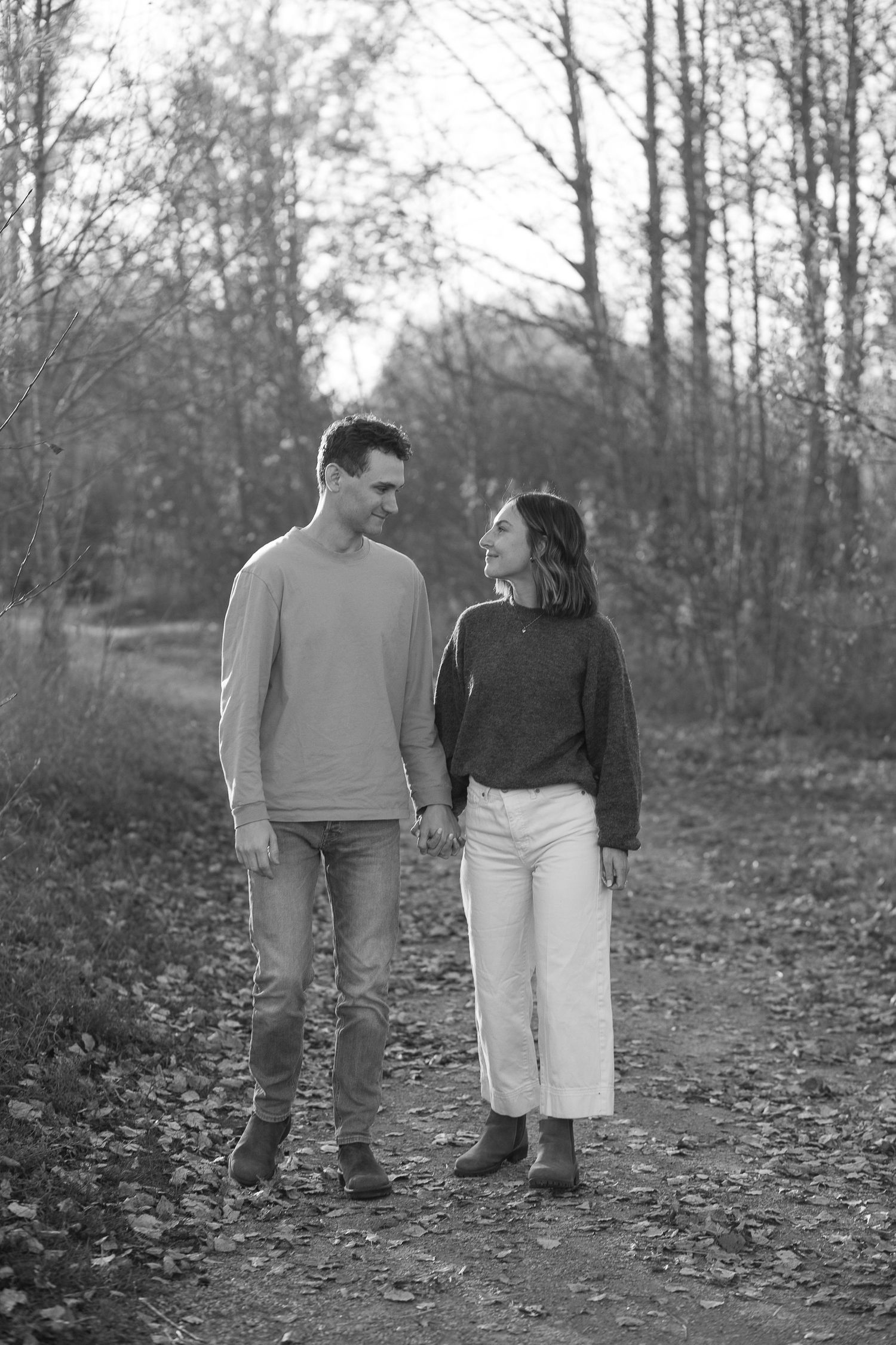 Black and white photo of a couple holding hands and smiling at each other on a leaf-covered autumn forest path.