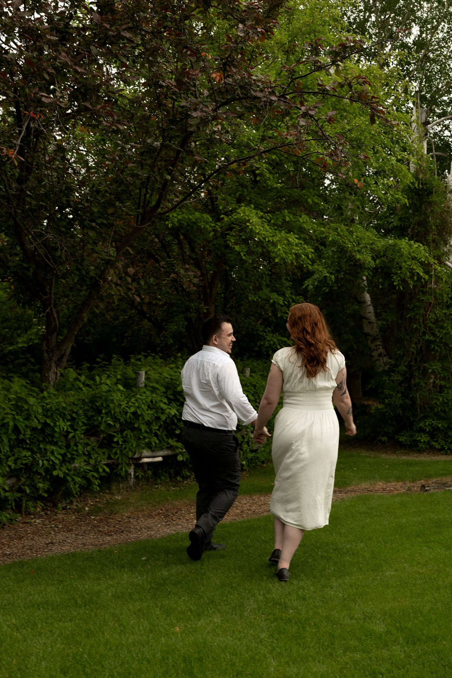 A couple in wedding attire walks hand-in-hand through a lush green garden, the bride in white and groom in black.