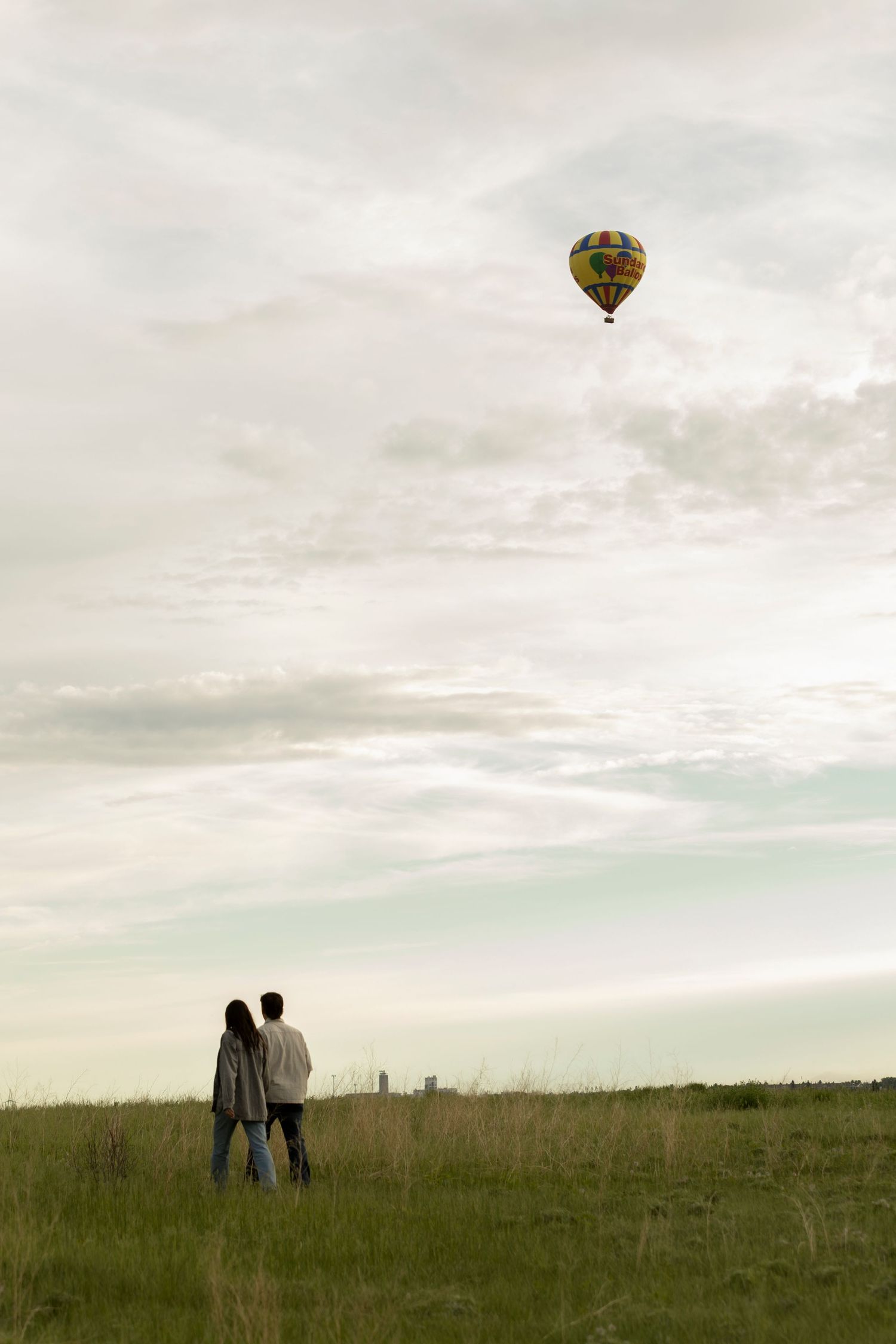 Couple standing in green field watching colorful hot air balloon float in cloudy sky.