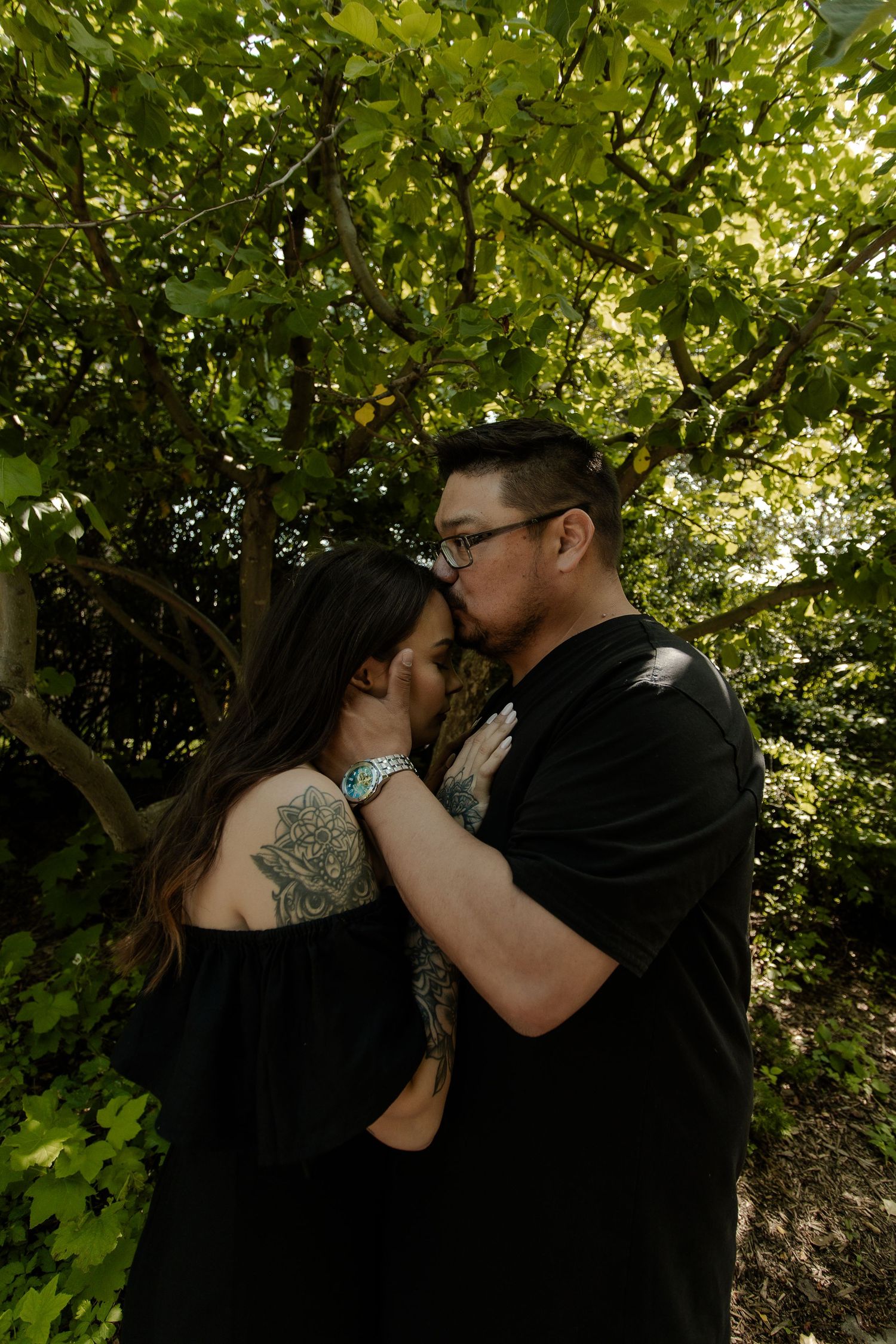 Couple sharing a tender kiss outdoors under lush green trees, both wearing black with visible tattoos.