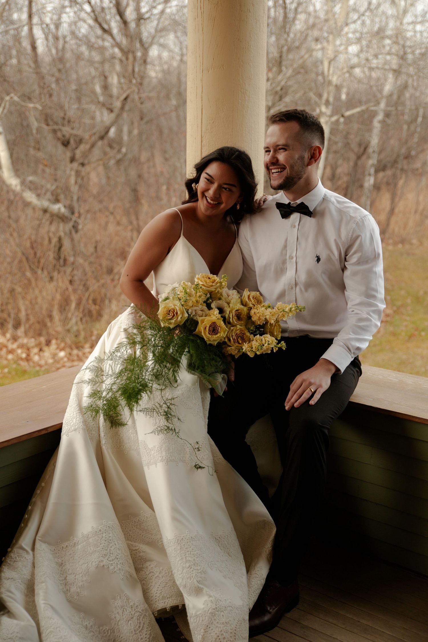 Bride and groom sitting together on a porch, smiling, bride holding yellow roses bouquet, groom in white shirt and bow tie.