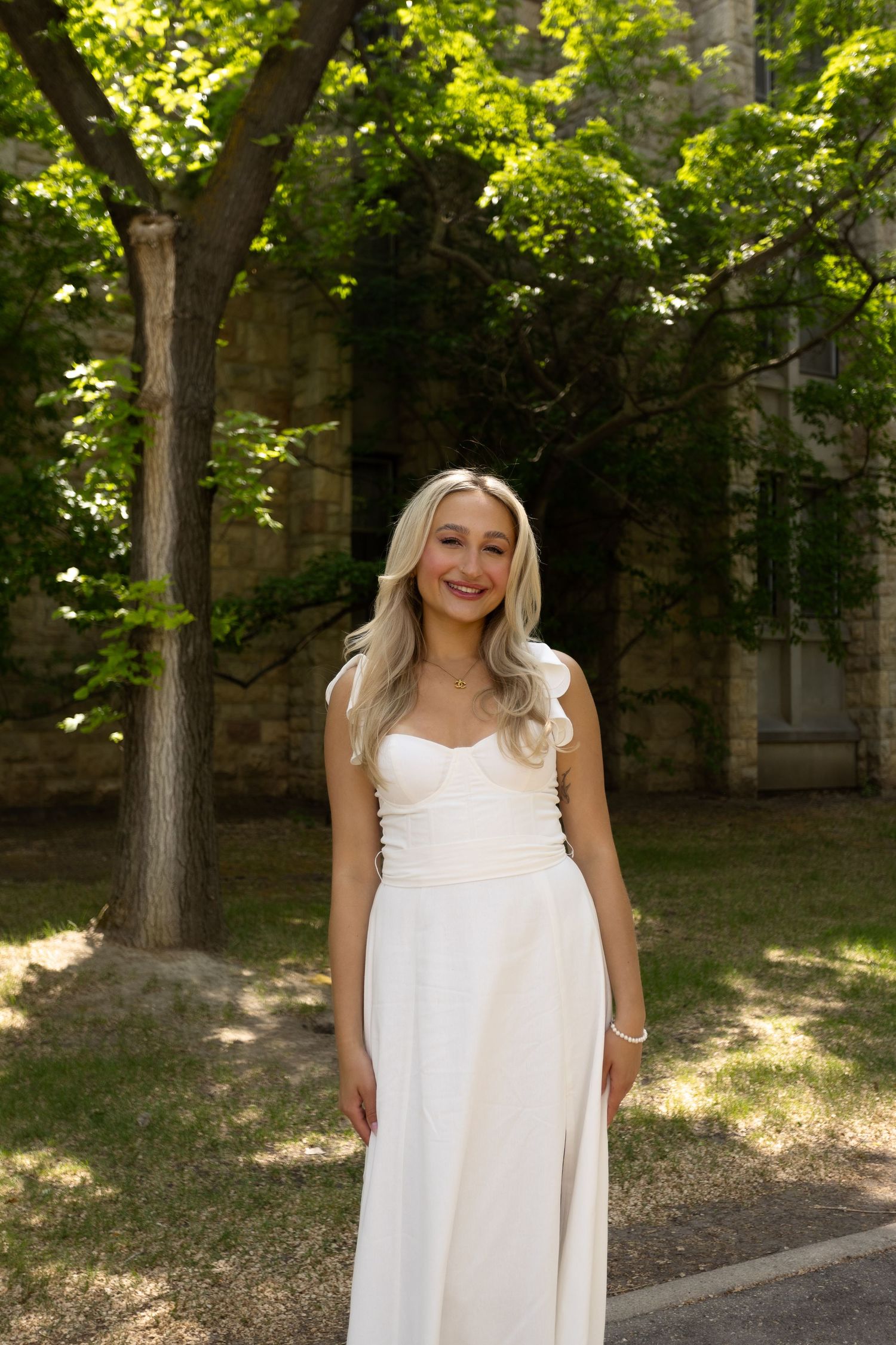 Blonde woman in elegant white one-shoulder gown smiling outdoors near stone building surrounded by green trees.