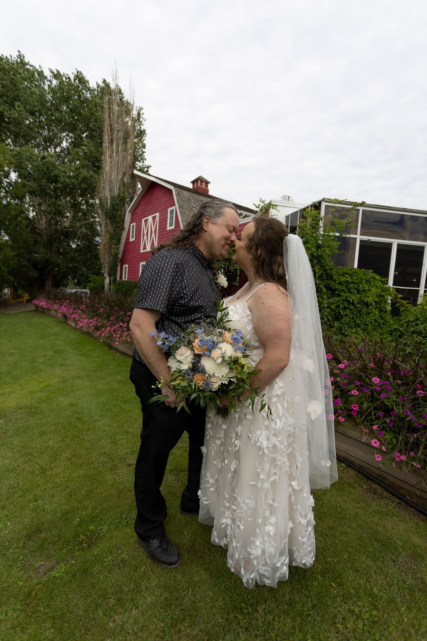Bride and groom share a kiss in front of a red barn, bride holding a blue and white bouquet in a lace wedding gown.