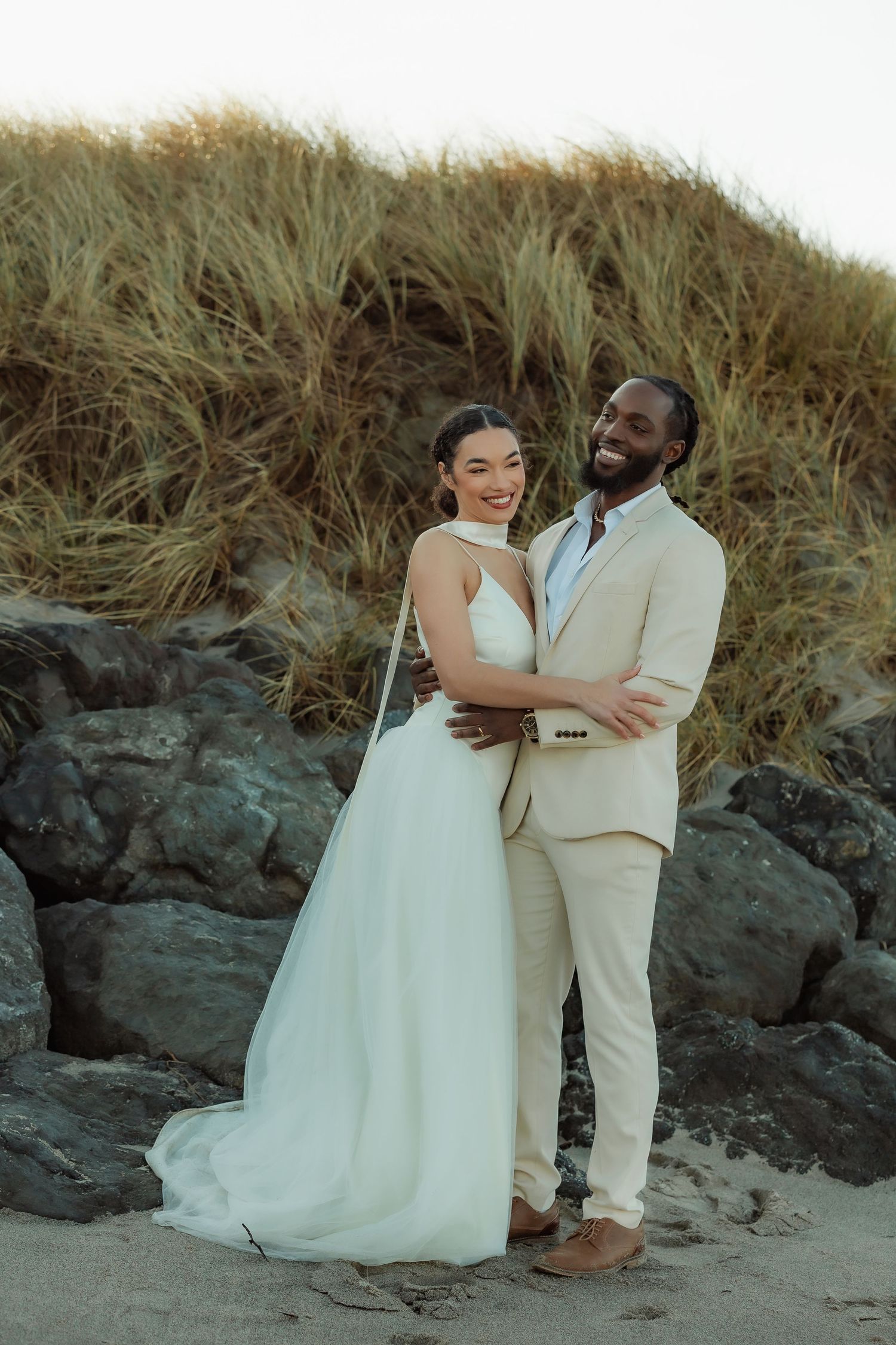 A couple in wedding attire embrace on a beach with sand dunes and tall grass in the background.