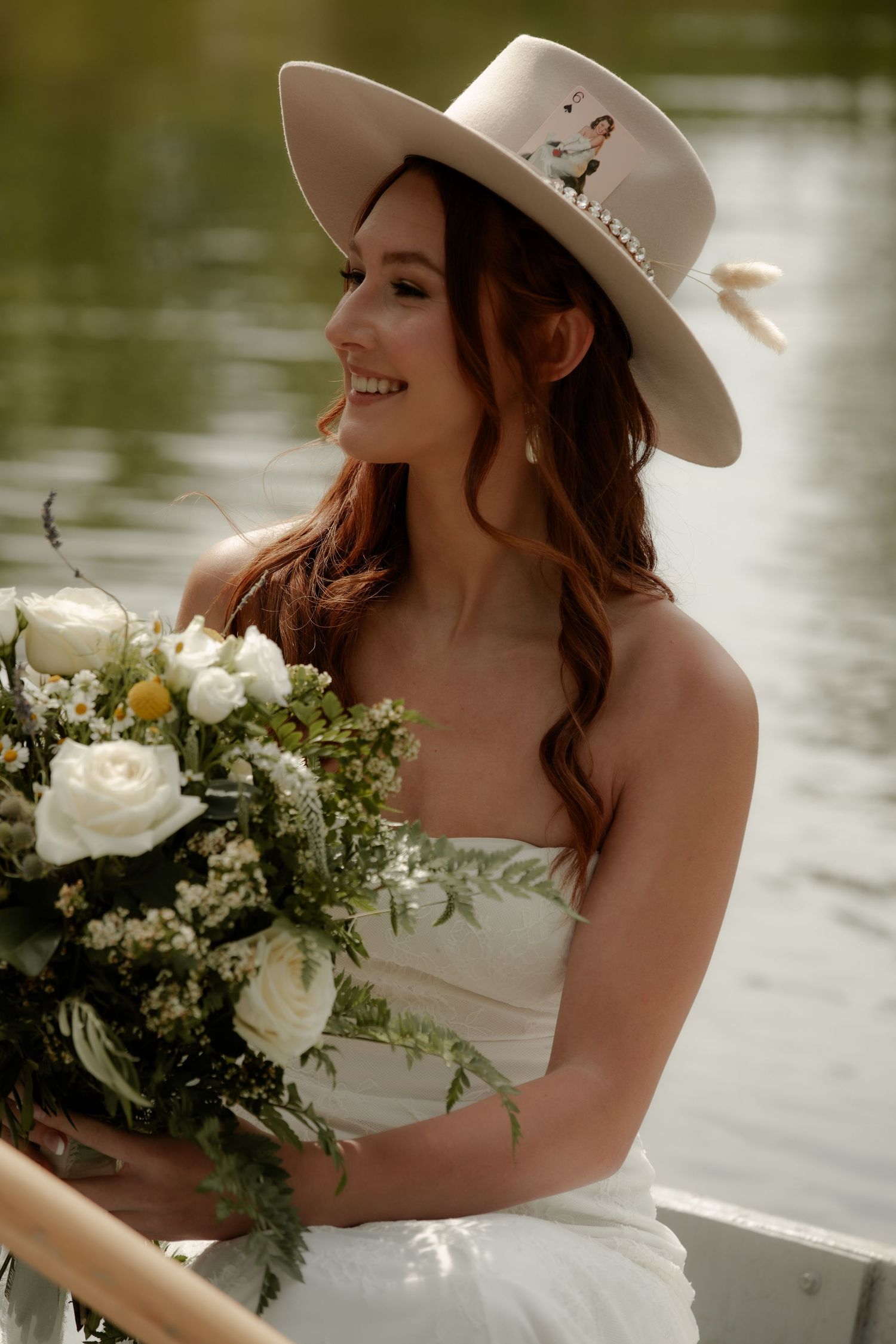 Person in white dress and western hat holding elegant floral bouquet by the water.