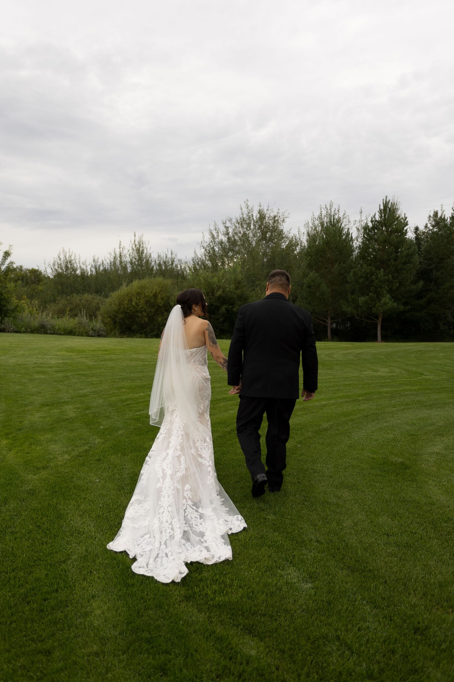 A couple in wedding attire walks across a lush green lawn with trees in the background at Barn at Wind's Edge in Saskatoon, Saskatchewan.