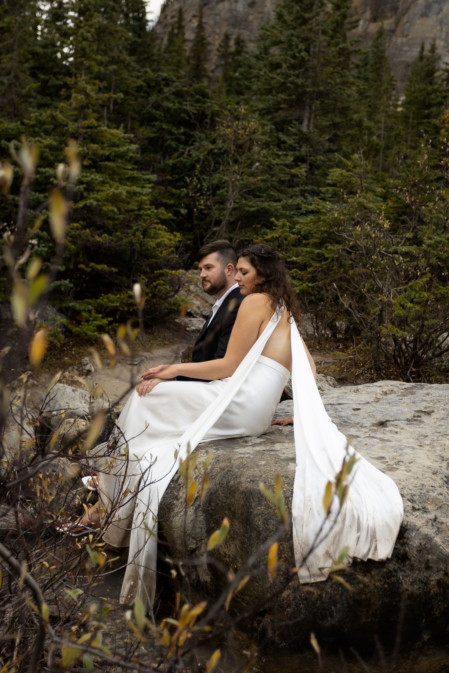 Bride and groom sitting on a rock surrounded by evergreen forest in a romantic outdoor wedding photo.