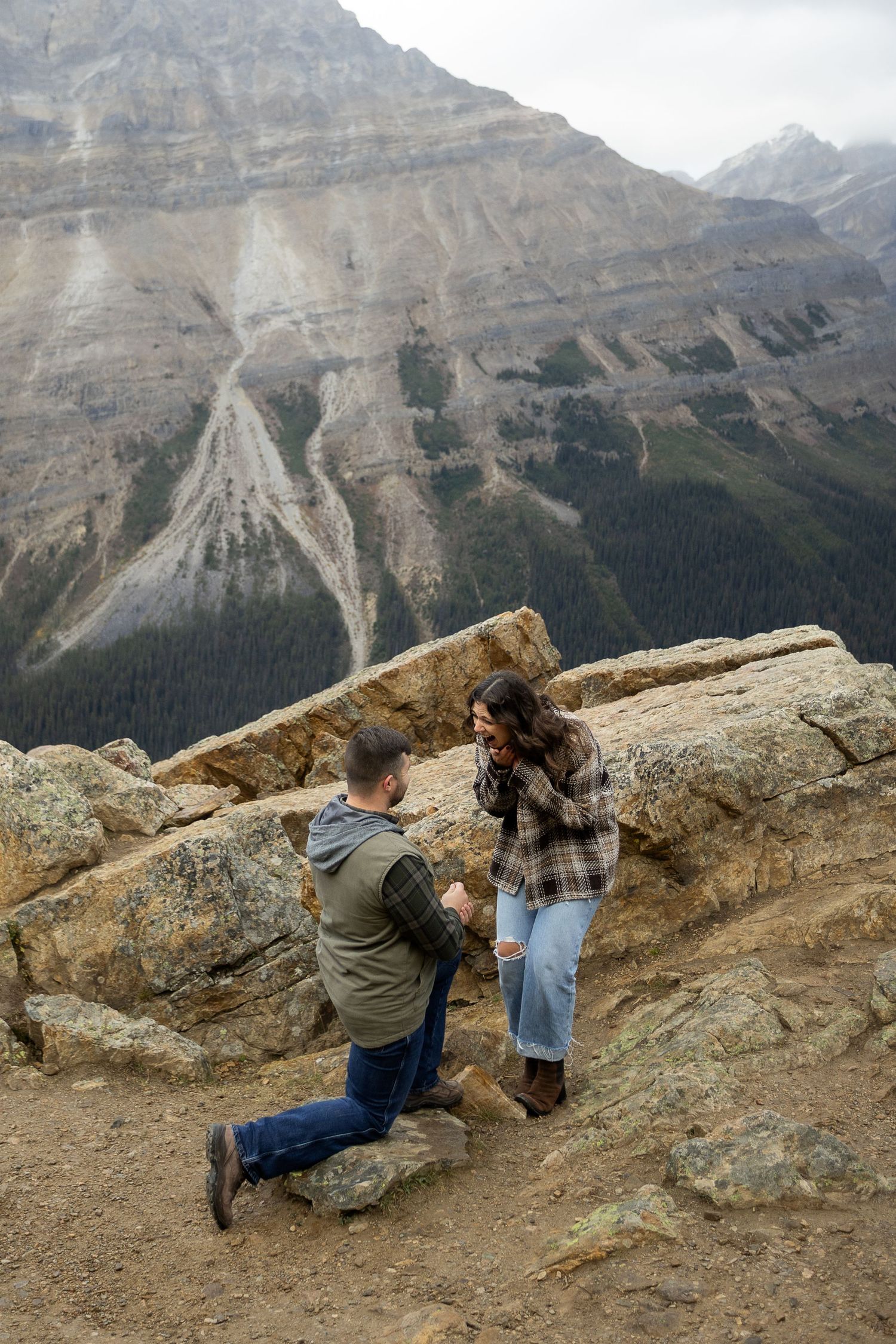 Man proposing on one knee to surprised woman on rocky mountain overlook with dramatic Canadian Rockies backdrop.