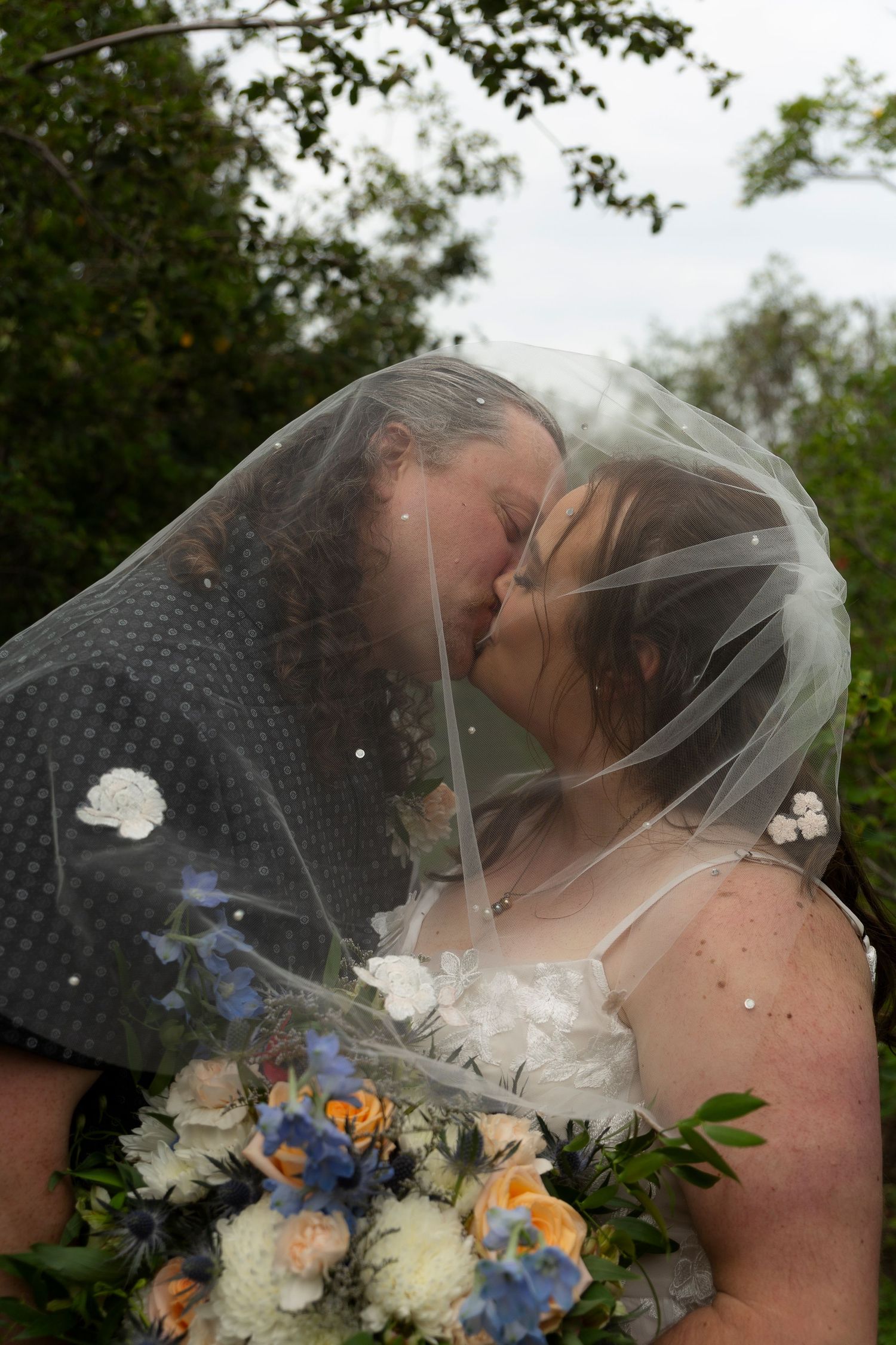 Two brides sharing a kiss under a wedding veil, one holding a bouquet of blue and white flowers outdoors.