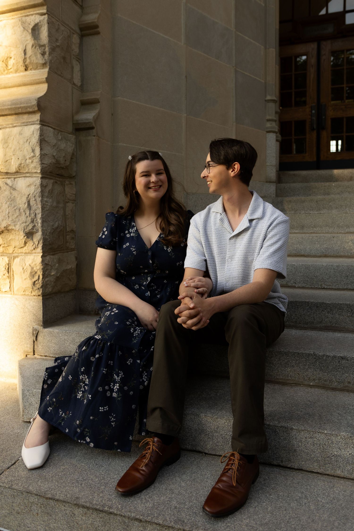 A smiling couple holds hands on stone steps outside a historic building, woman in floral dress, man in white shirt.