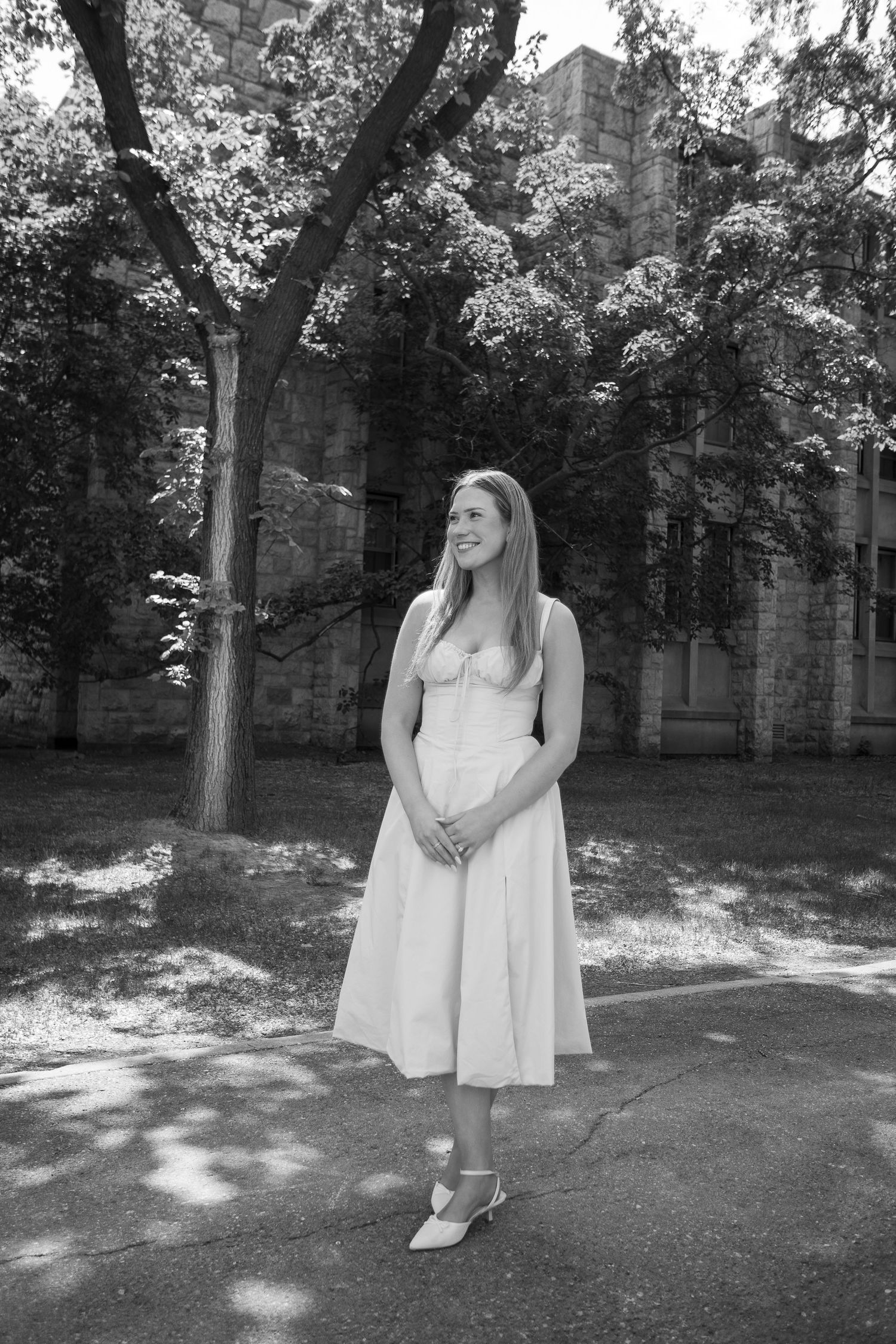 Young woman in white midi dress smiling outdoors near historic stone building with trees in black and white photo.