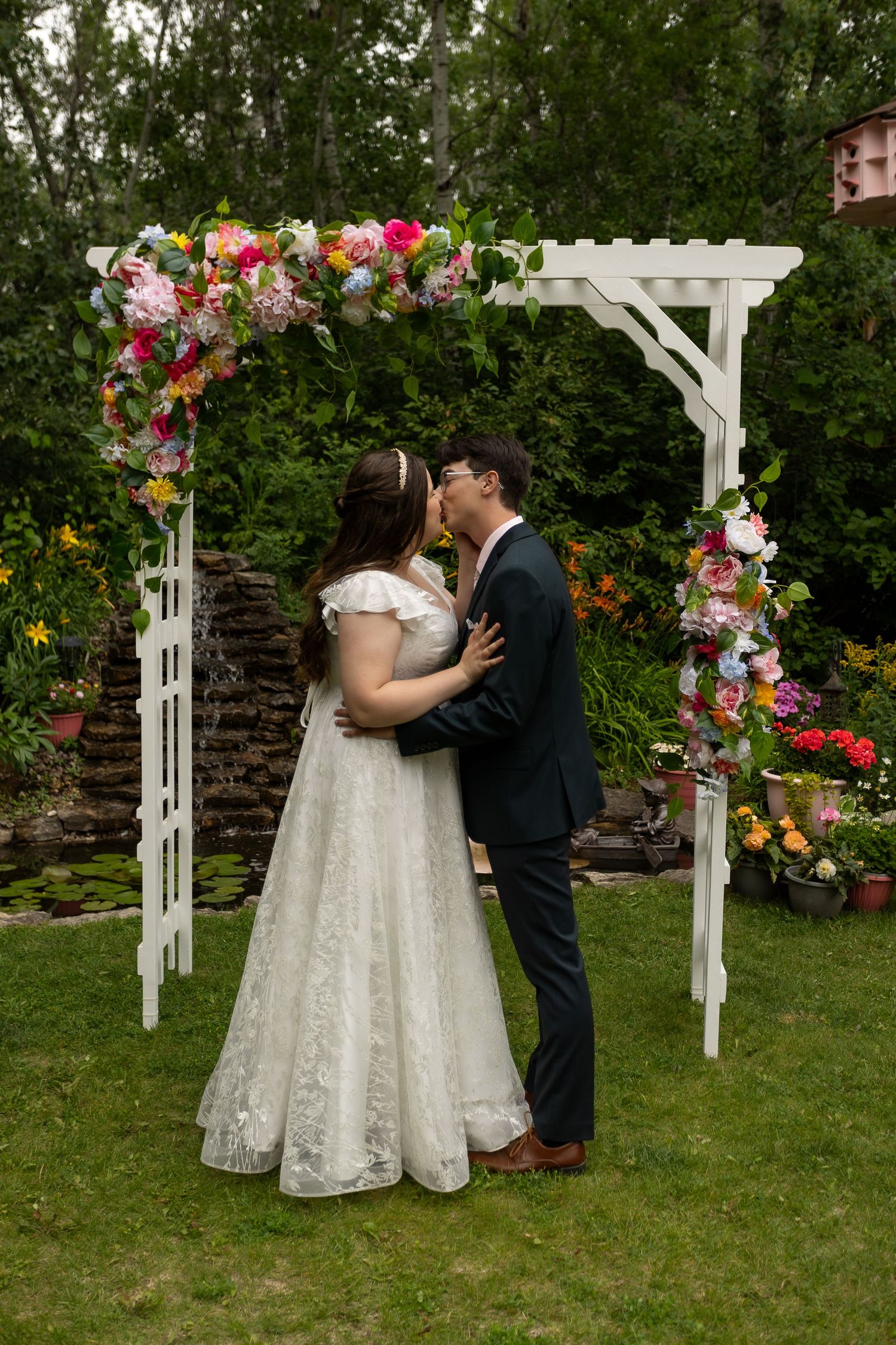 Newlyweds kiss under a white garden arch decorated with colorful flowers.