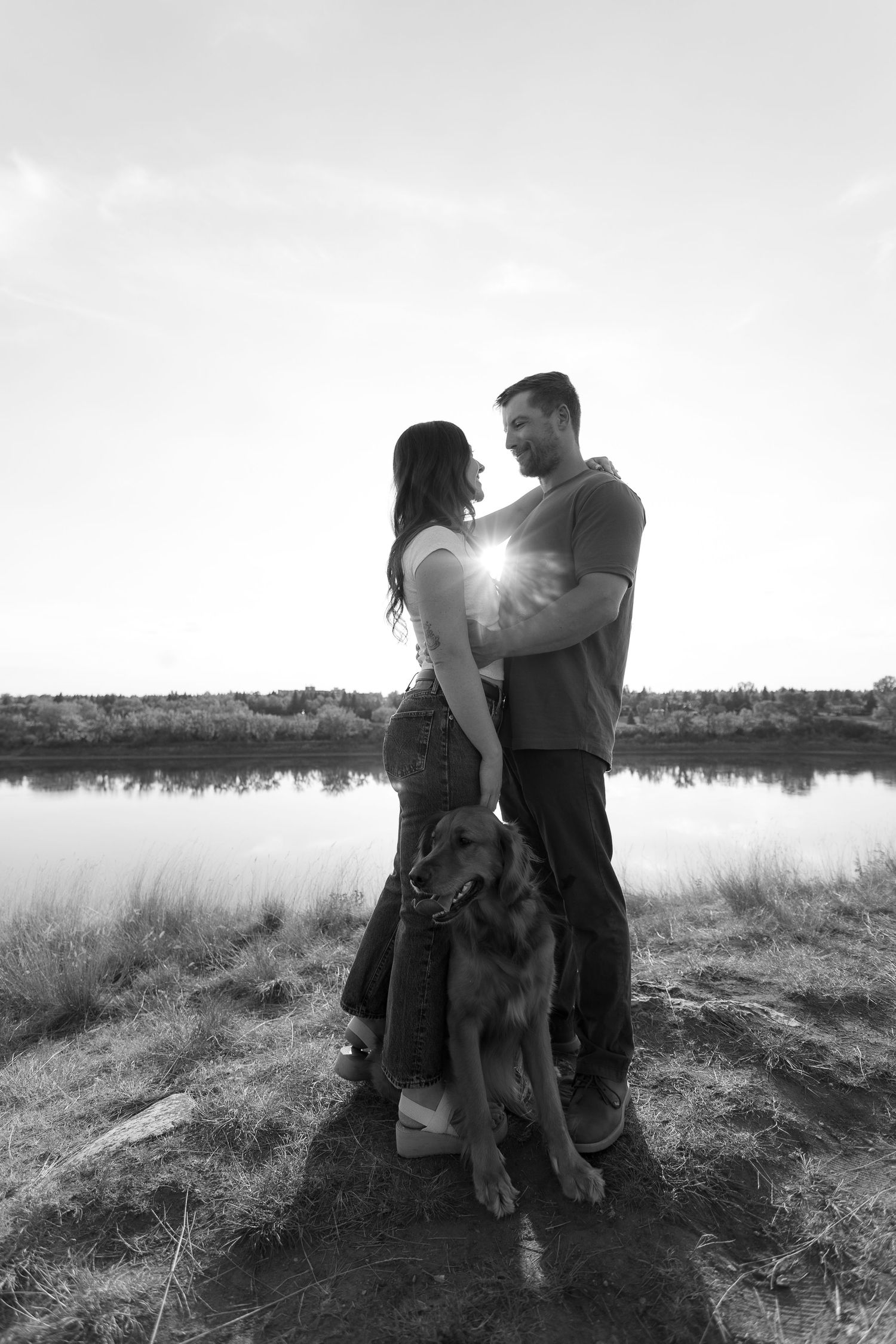 A black and white portrait of a couple standing by a lake with their dog against a cloudy sky at Crocus Prairies in Saskatoon, Saskatchewan.