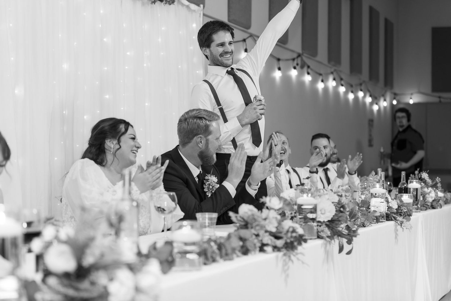 Black and white wedding reception photo of a groomsman giving a toast while guests laugh and applaud at a decorated head table.