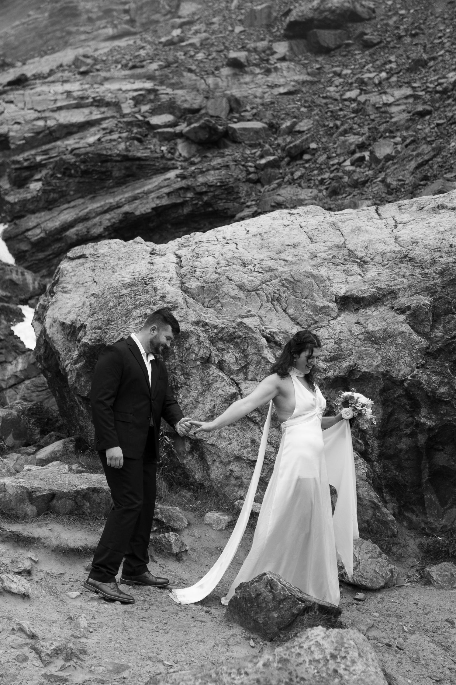 Black and white photo of bride and groom holding hands on rocky terrain, bride holding bouquet in flowing wedding gown.