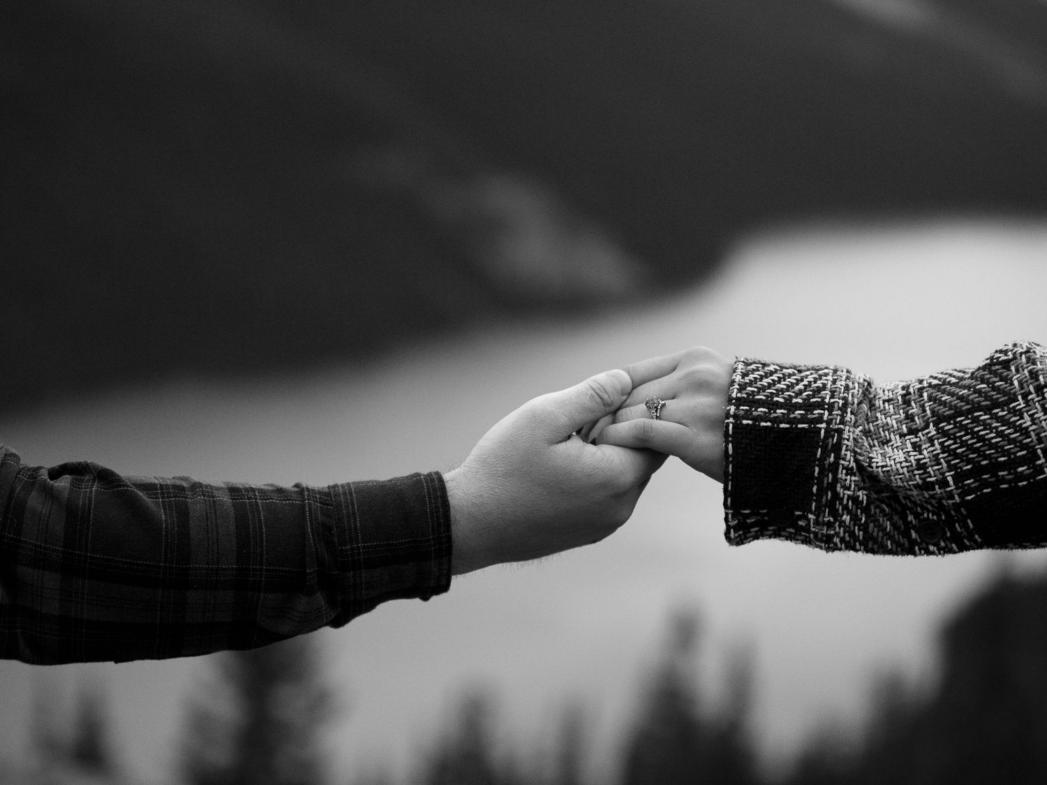 Black and white photo of two people gently holding hands outdoors with a blurred mountain lake background.