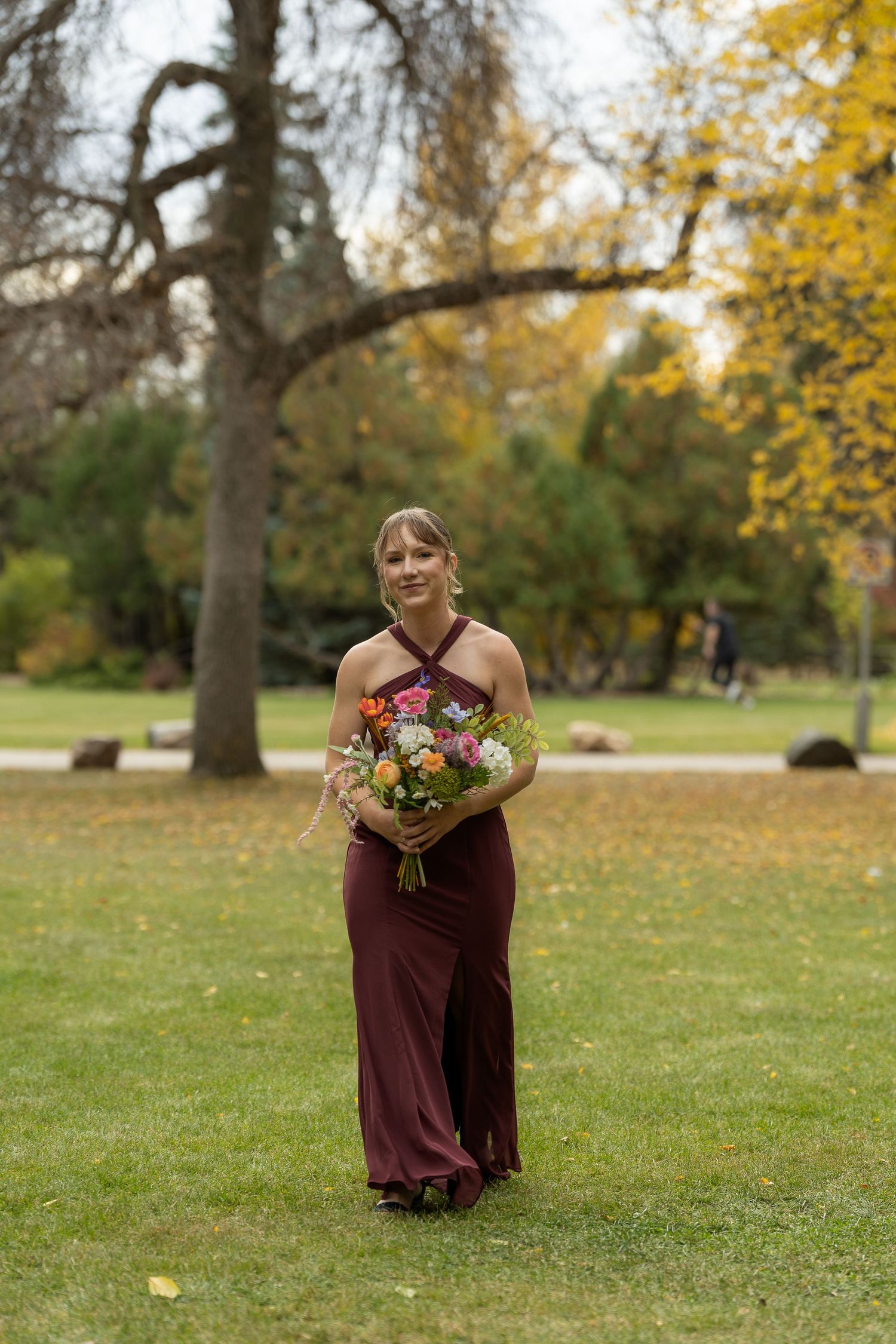 Bridesmaid in burgundy dress holding autumn bouquet in park setting.