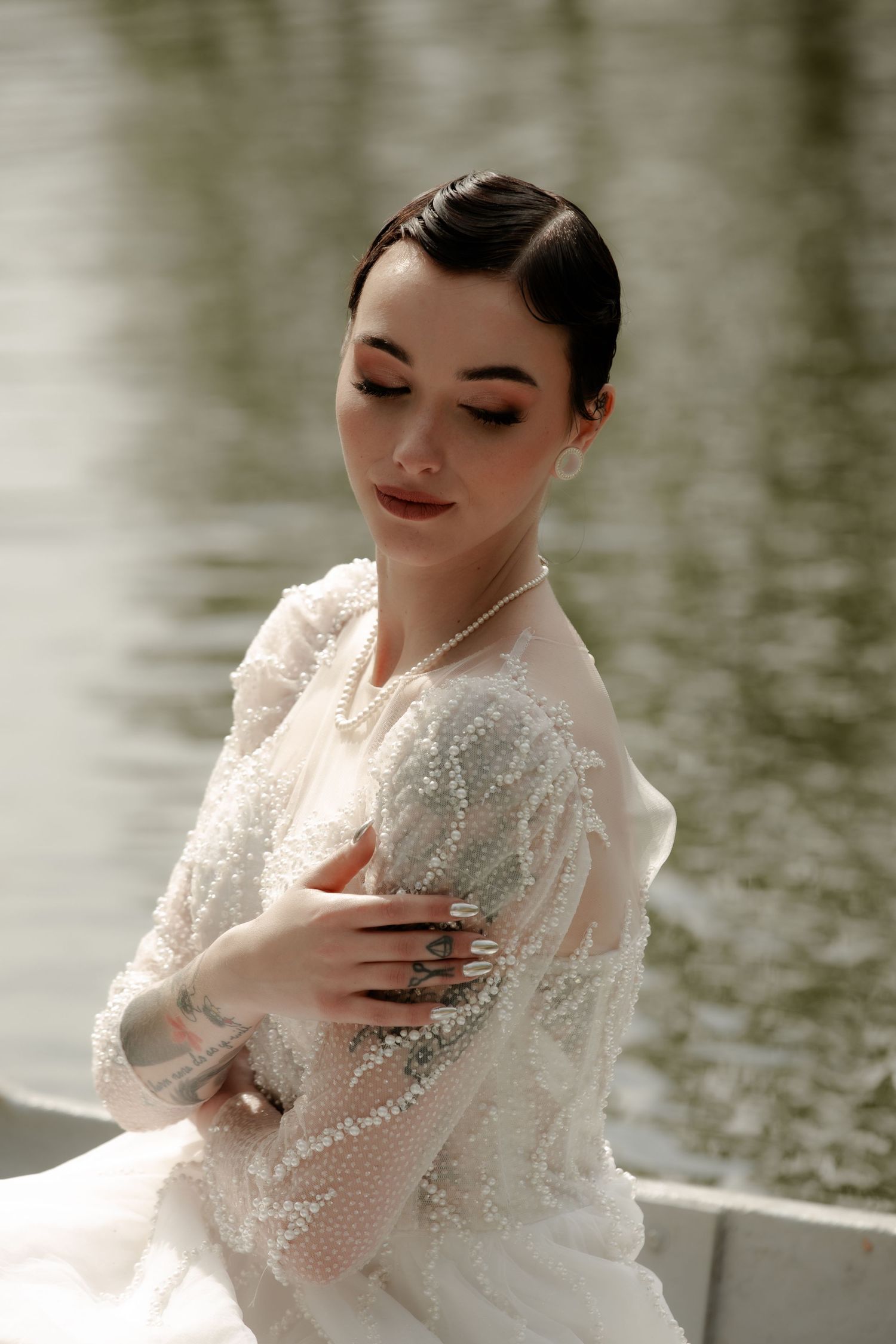 A bride in a sparkly white gown poses elegantly by a lake.