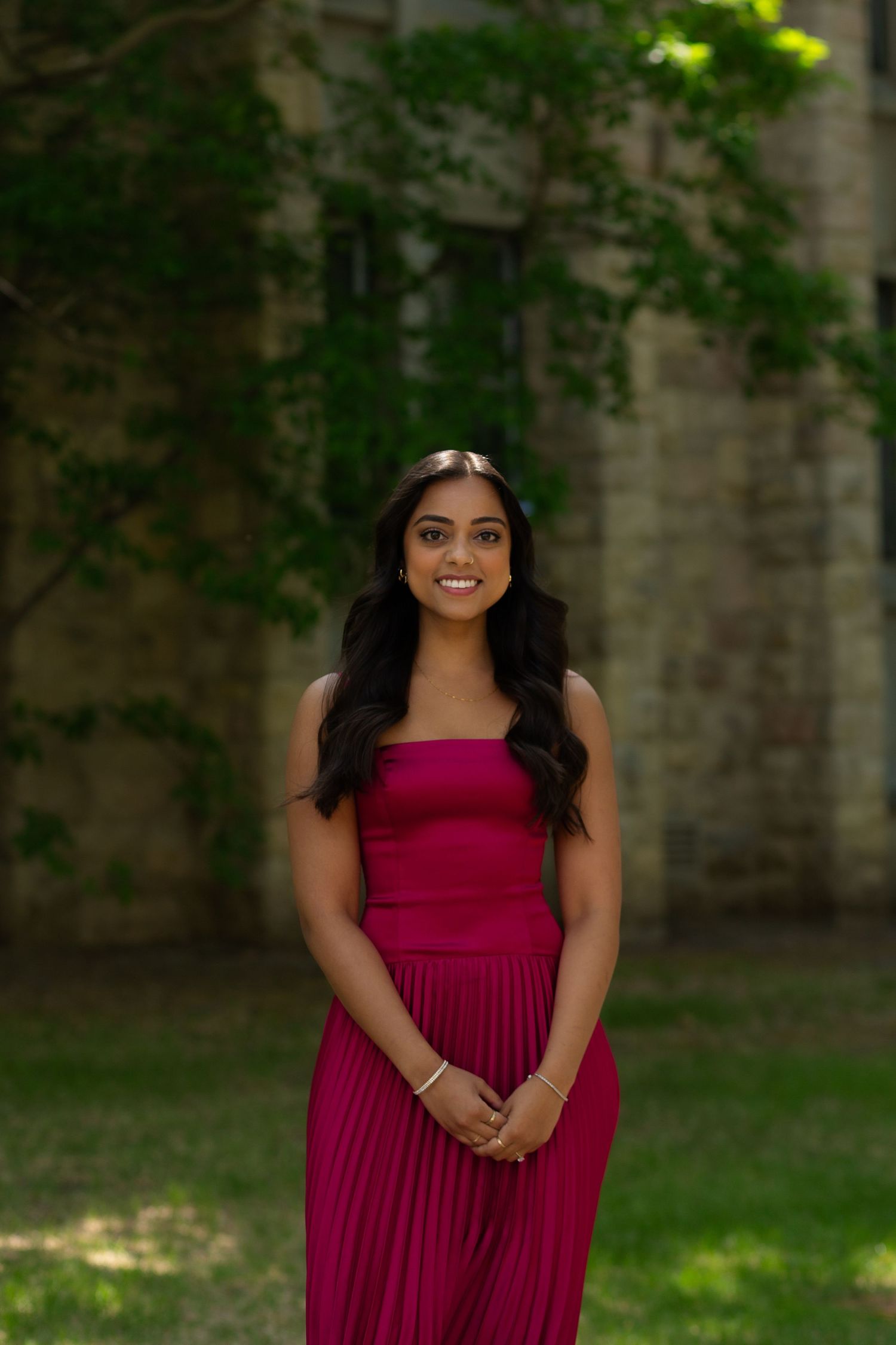 Young woman in magenta strapless pleated dress smiling outdoors near stone building with lush green trees.