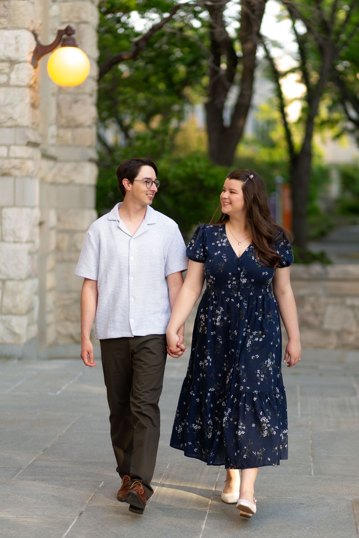 A smiling couple walks hand-in-hand outdoors, man in white shirt and woman in navy floral dress, with stone walls and trees.