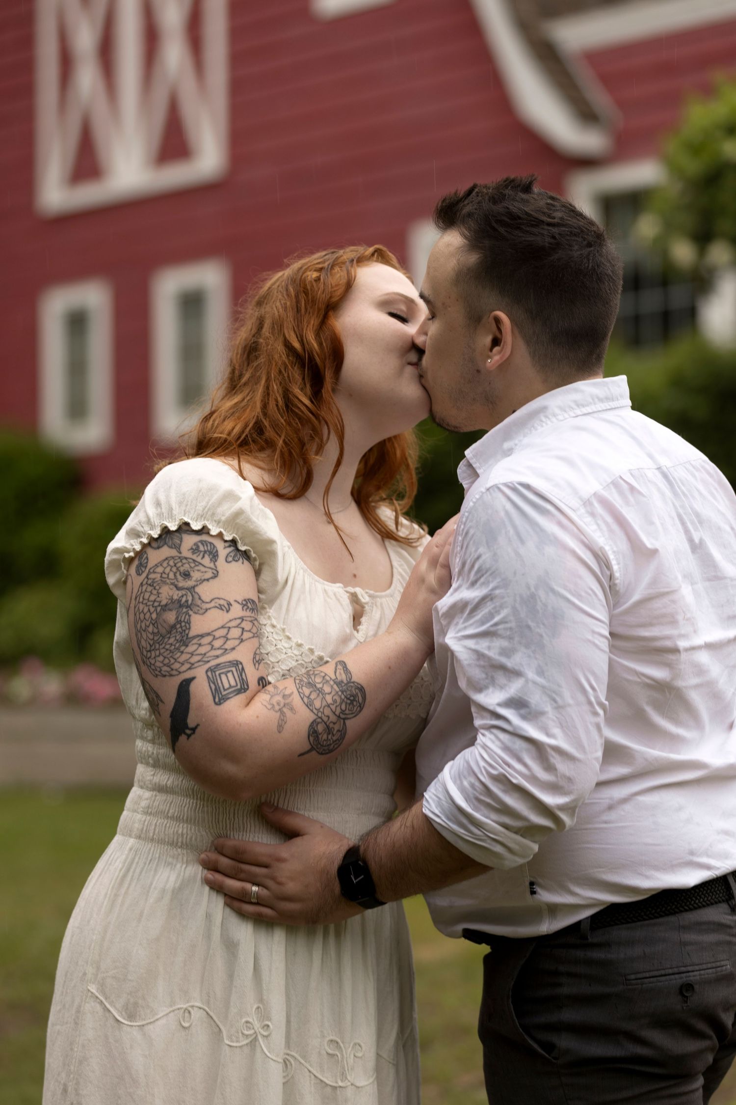 A tattooed redhead woman in a white dress kisses a man in front of a red barn.