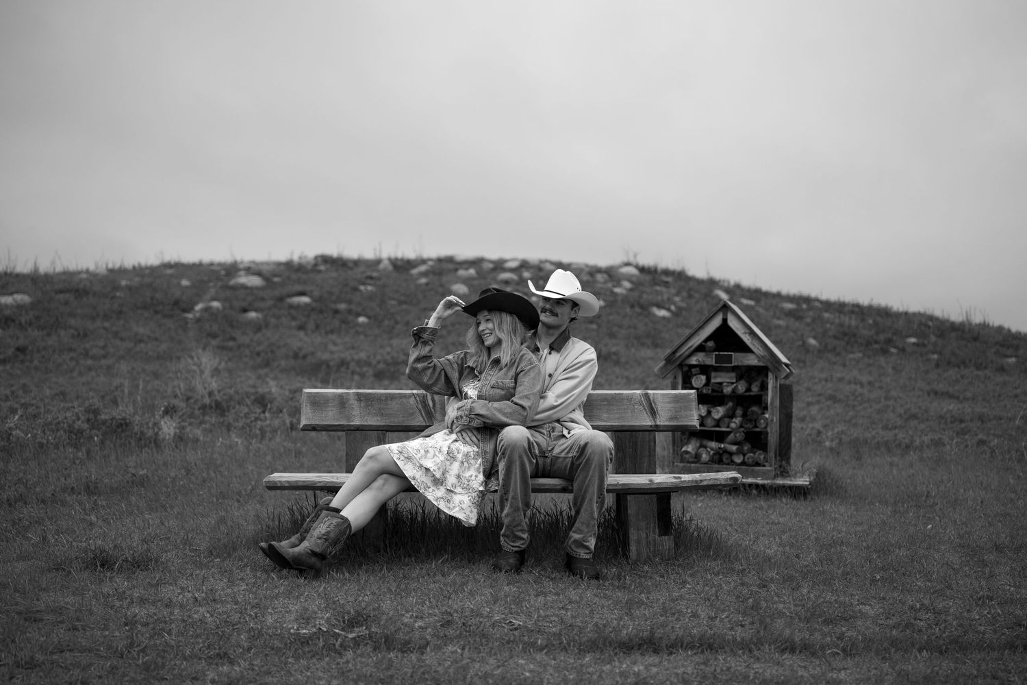 Black and white photo of a couple in cowboy hats sitting on a bench in a grassy field on an overcast day.