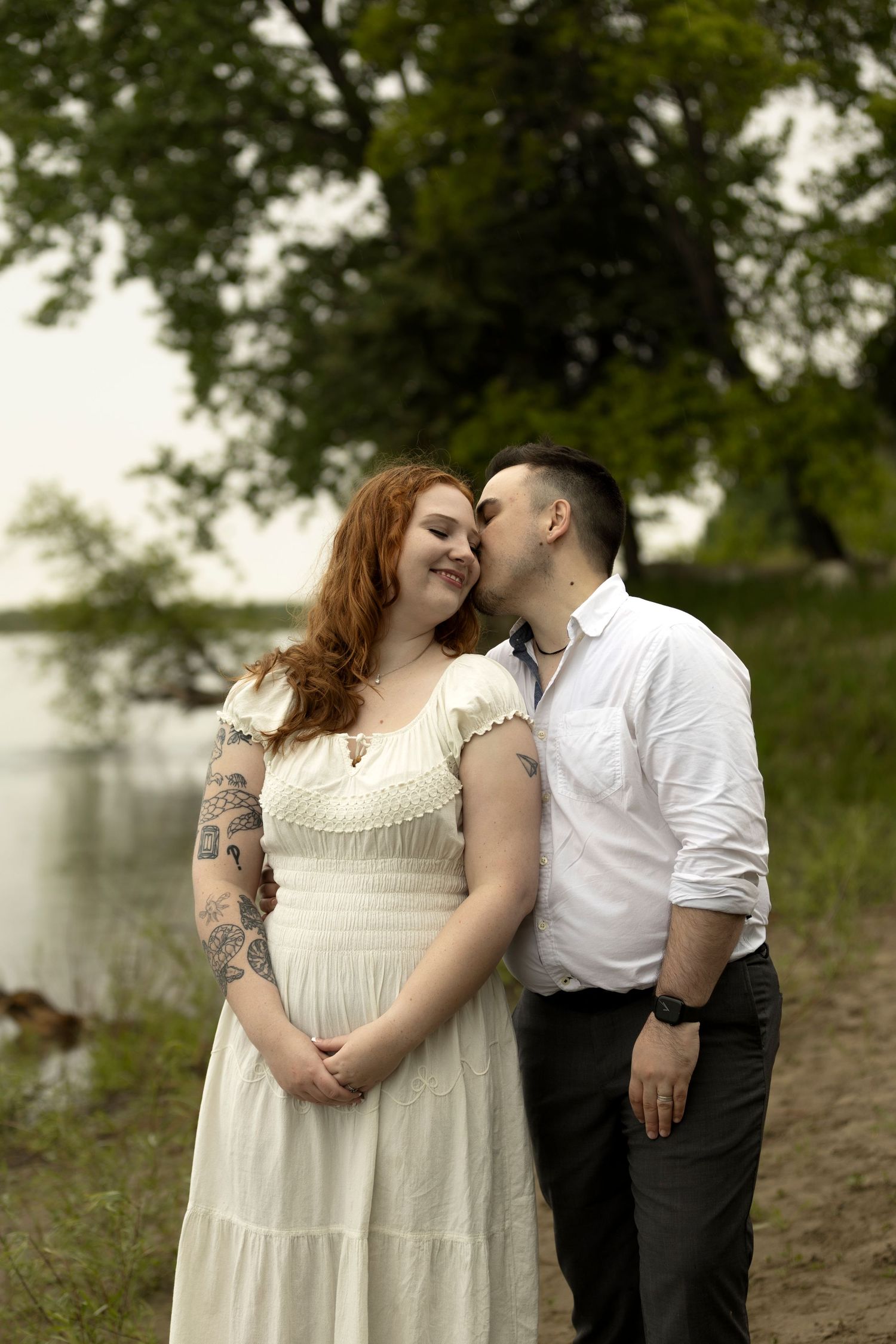 A man kisses a smiling redheaded woman with tattoos in a white dress outdoors near a lake.