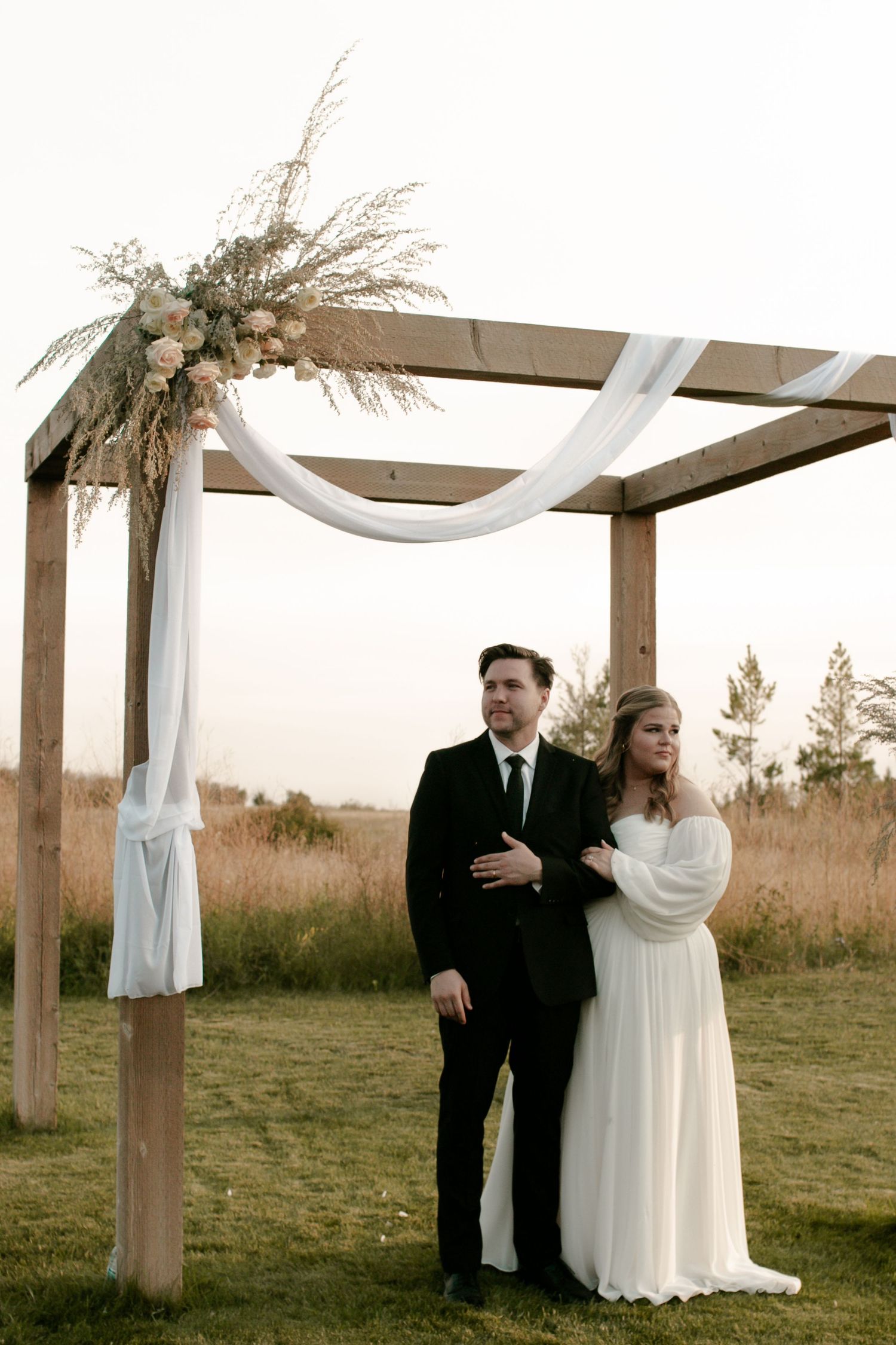 Newlyweds pose under a wooden pergola adorned with dried flowers and flowing white fabric at sunset.
