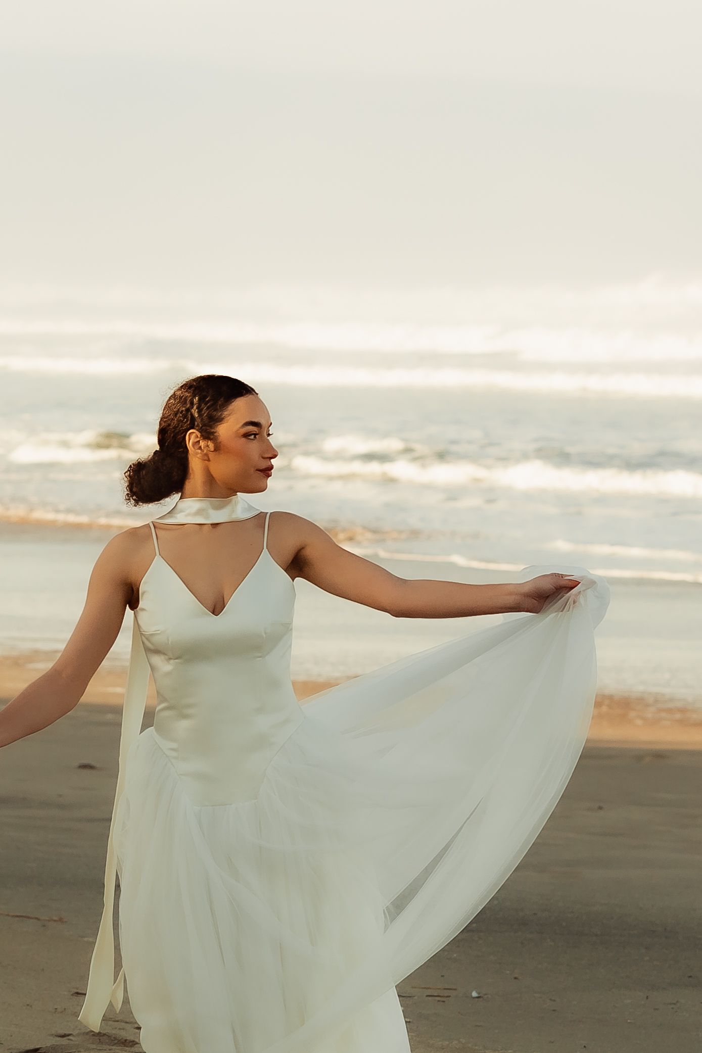 Bride in flowing white dress twirls on beach at sunset.