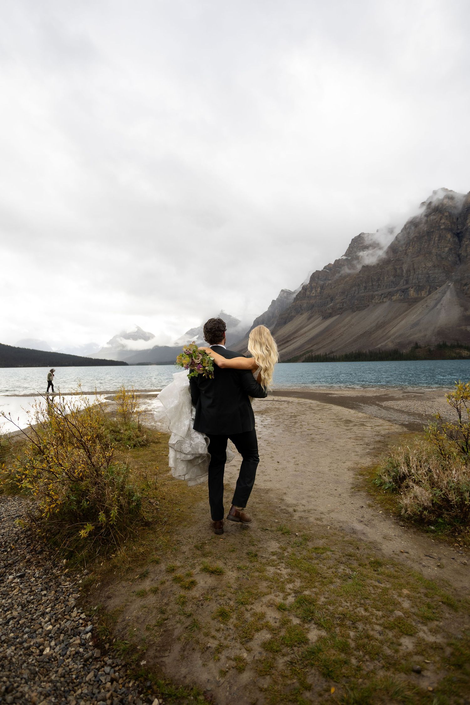 Bride and groom walking hand in hand toward a glacial lake with dramatic mountain backdrop on their wedding day.
