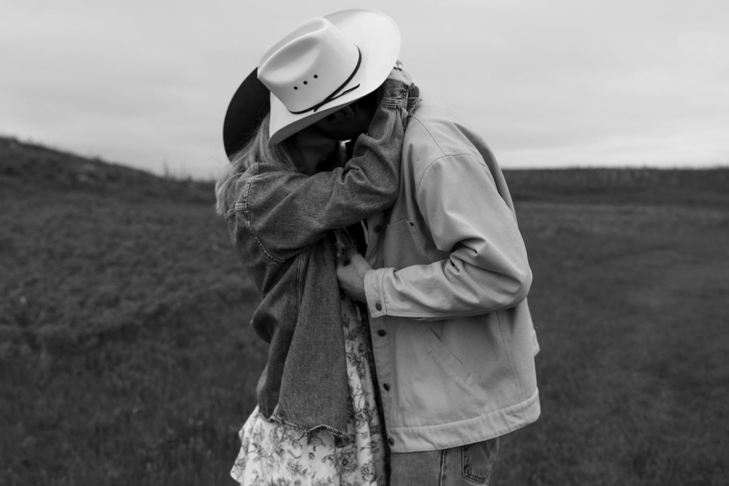 Black and white photo of a couple kissing in a field, man wearing a cowboy hat and jacket outdoors.