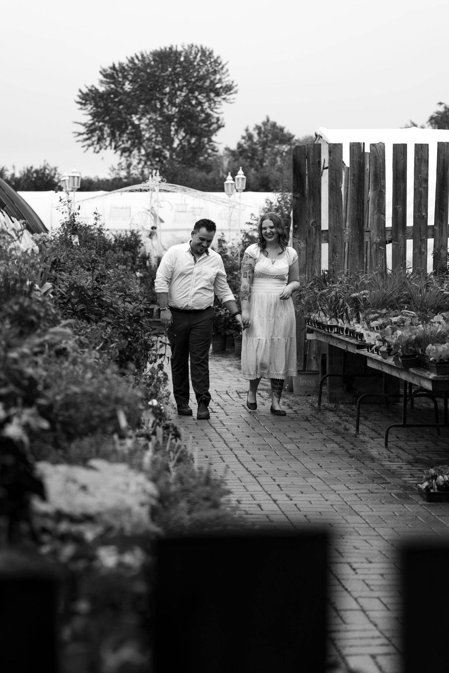 Couple walking hand in hand through a lush garden nursery pathway in black and white photography.