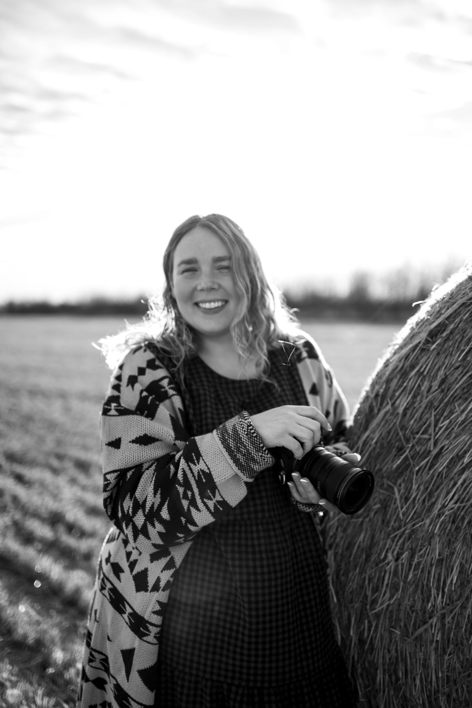 Black and white photo of a smiling woman holding a camera leaning against a hay bale in a rural field.