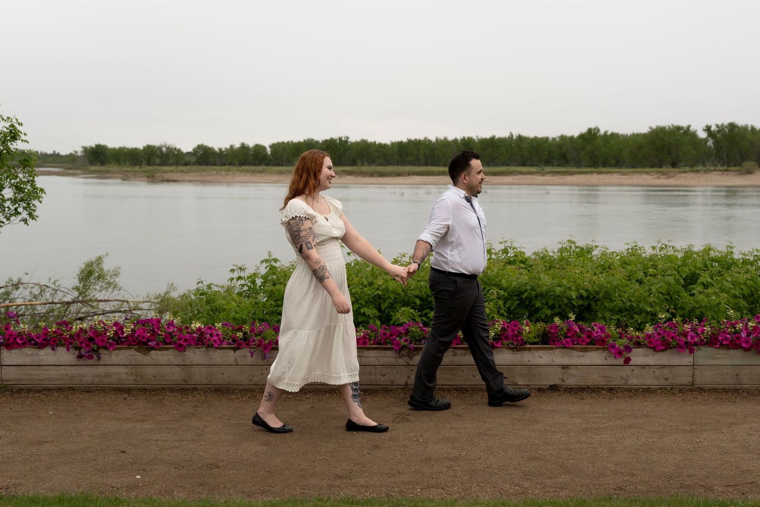 Couple holding hands walking along a scenic riverside path with pink flowers and trees in the background.