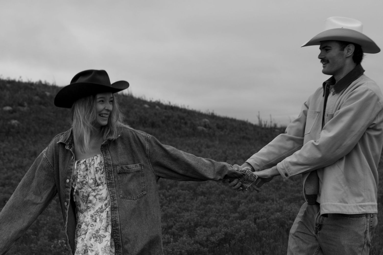 Black and white photo of a couple in cowboy hats holding hands and smiling outdoors on a hillside.