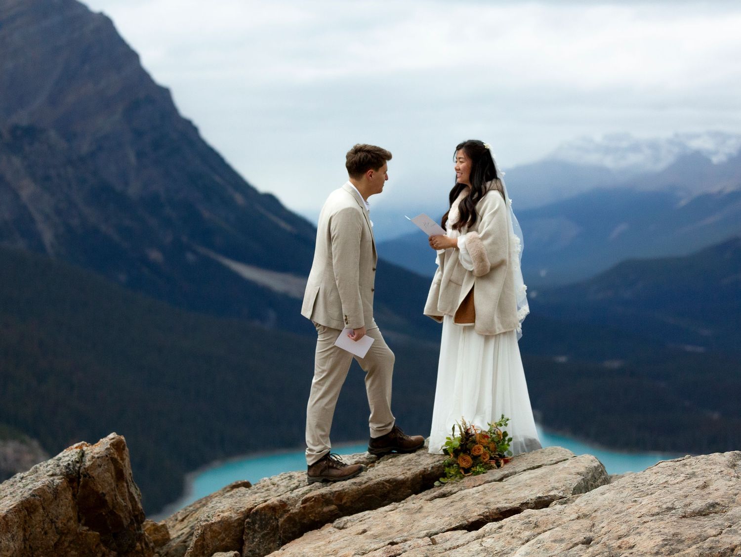 A couple in wedding attire stands on a mountain cliff overlooking a turquoise lake and dramatic mountain peaks at Petyo Lake.