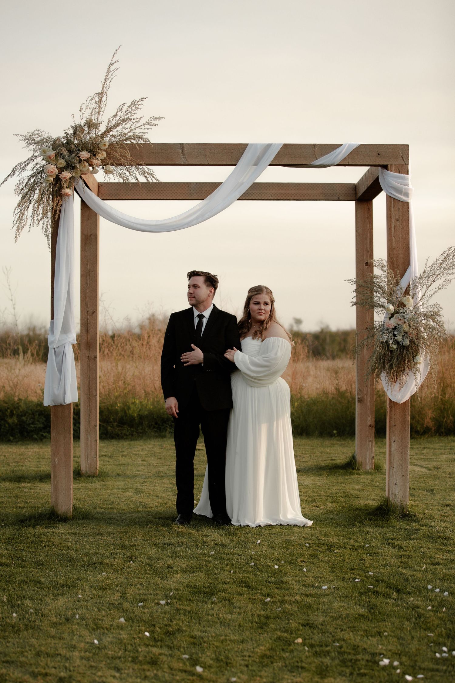 A wedding couple stands beneath a rustic wooden arch decorated with pampas grass and white fabric in an outdoor setting.