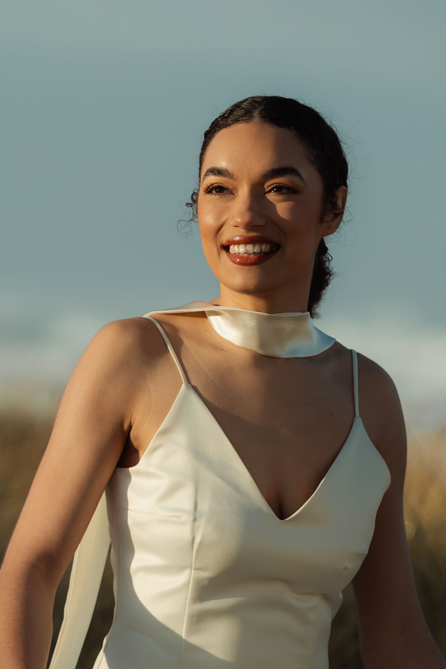 Bride in elegant white slip dress smiles in golden sunset light.