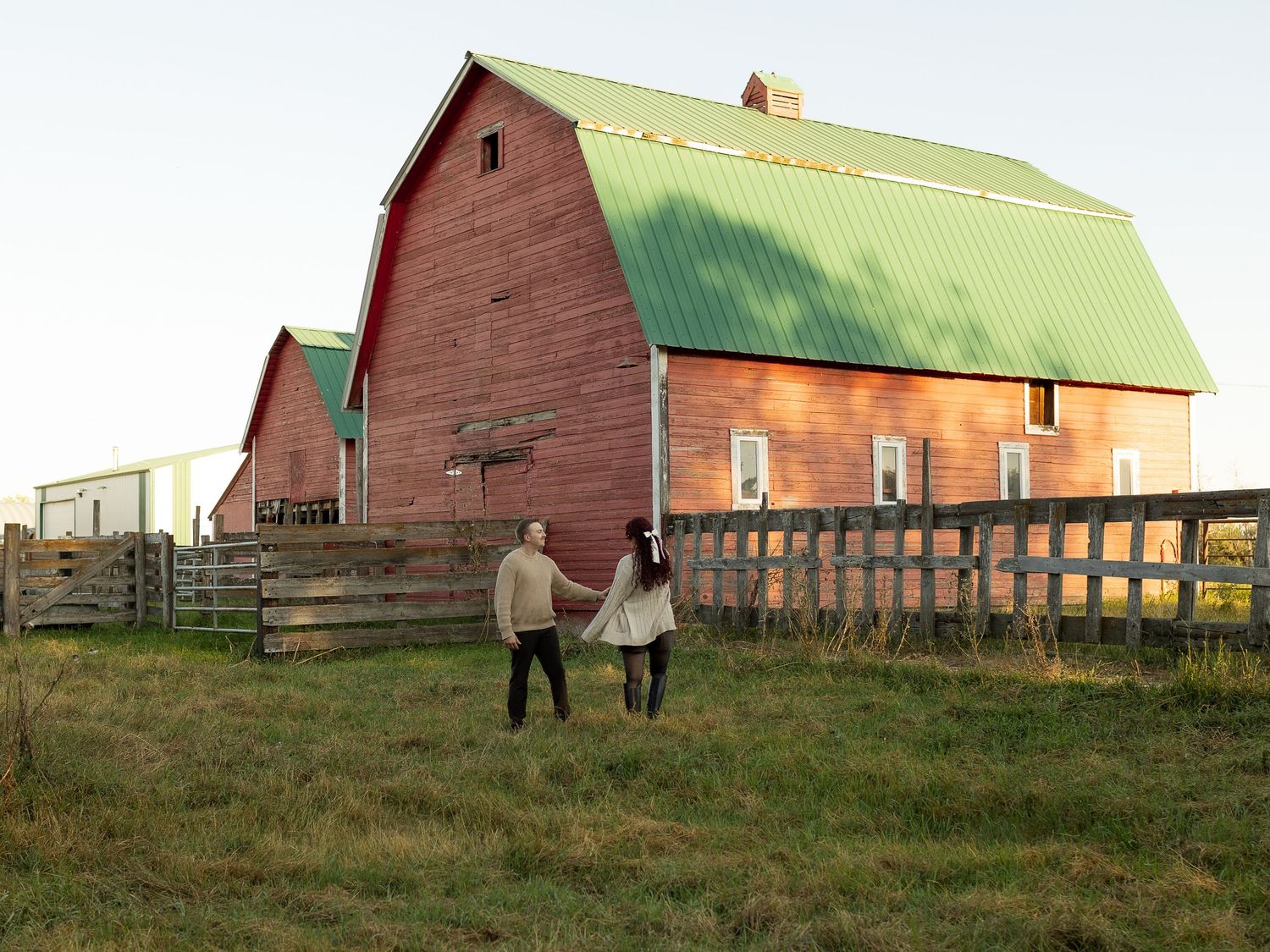 A couple stands together in front of a red barn during golden hour on a farm in the Canadian prairies.