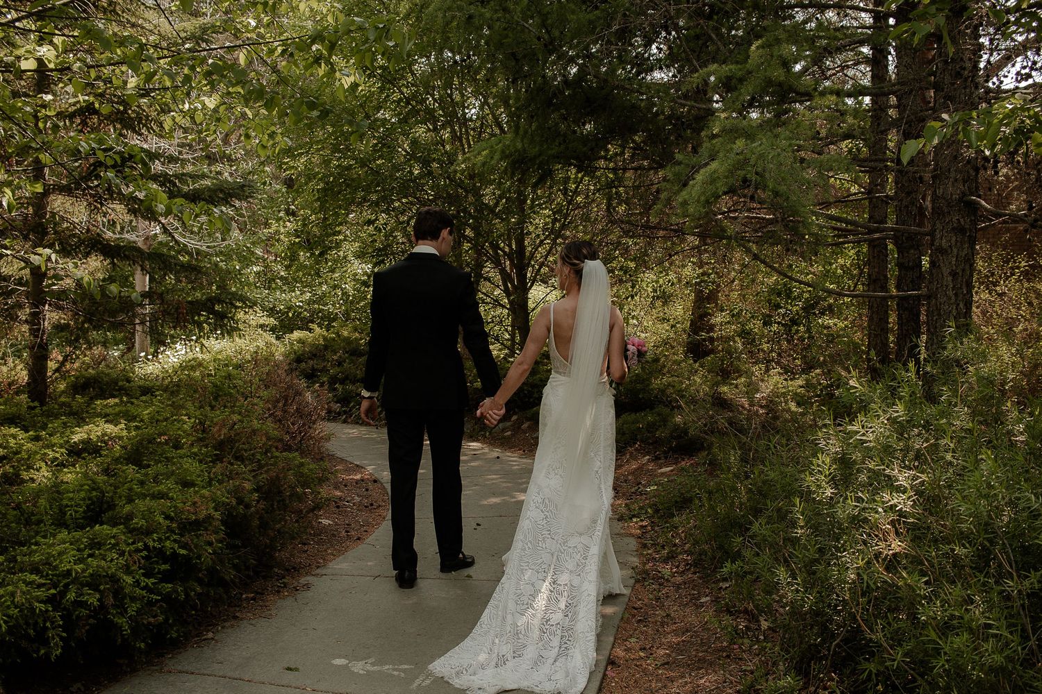 A couple in wedding attire walks along a scenic forest path surrounded by lush greenery and tall trees.