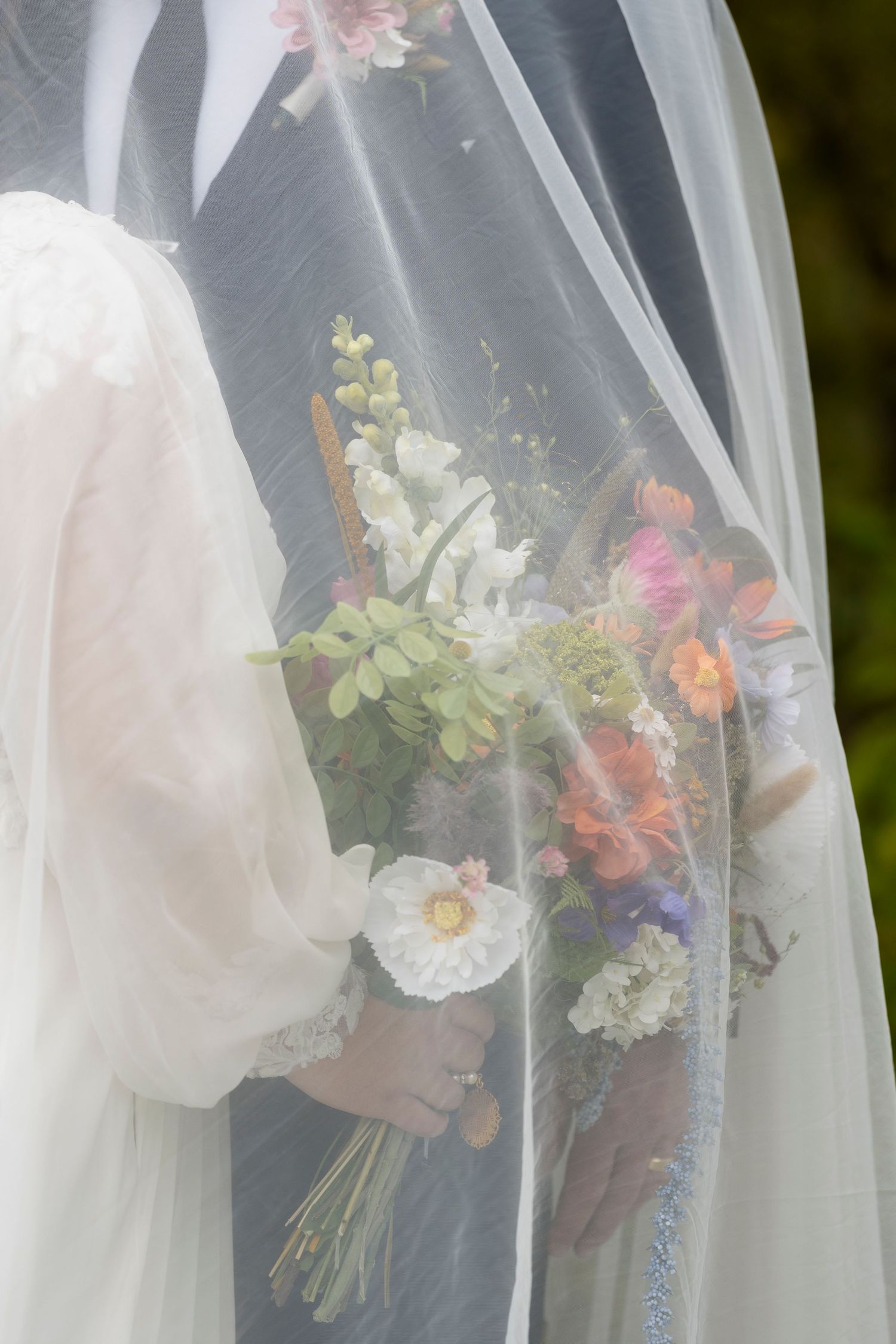 Bride holding a colorful wildflower bouquet, partially veiled in white tulle at a wedding ceremony.