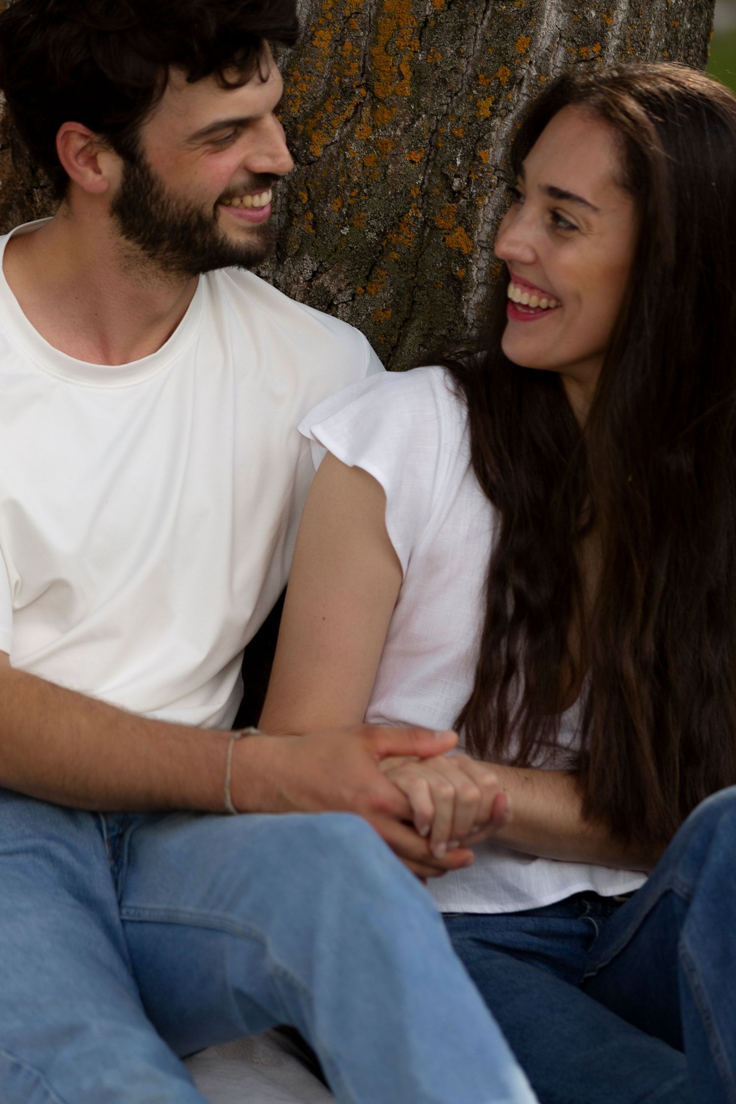 Couple in white t-shirts and jeans sharing laughs while leaning against tree trunk.