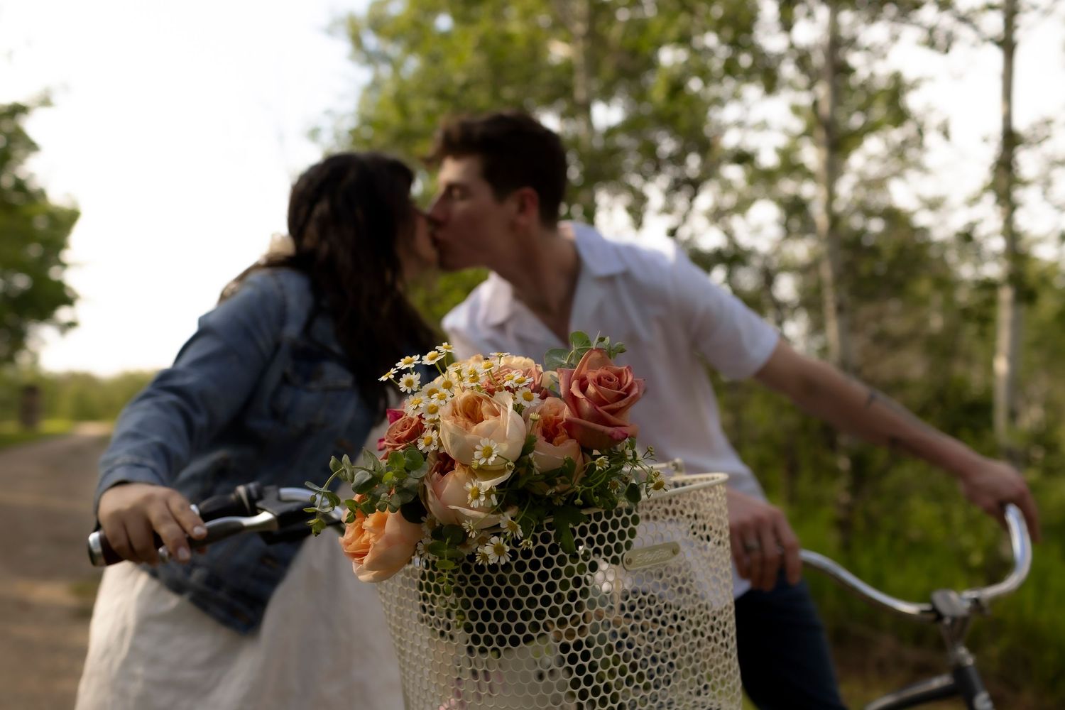 Couple kissing on bicycles with a white basket of orange and cream roses in the foreground on a tree-lined path.