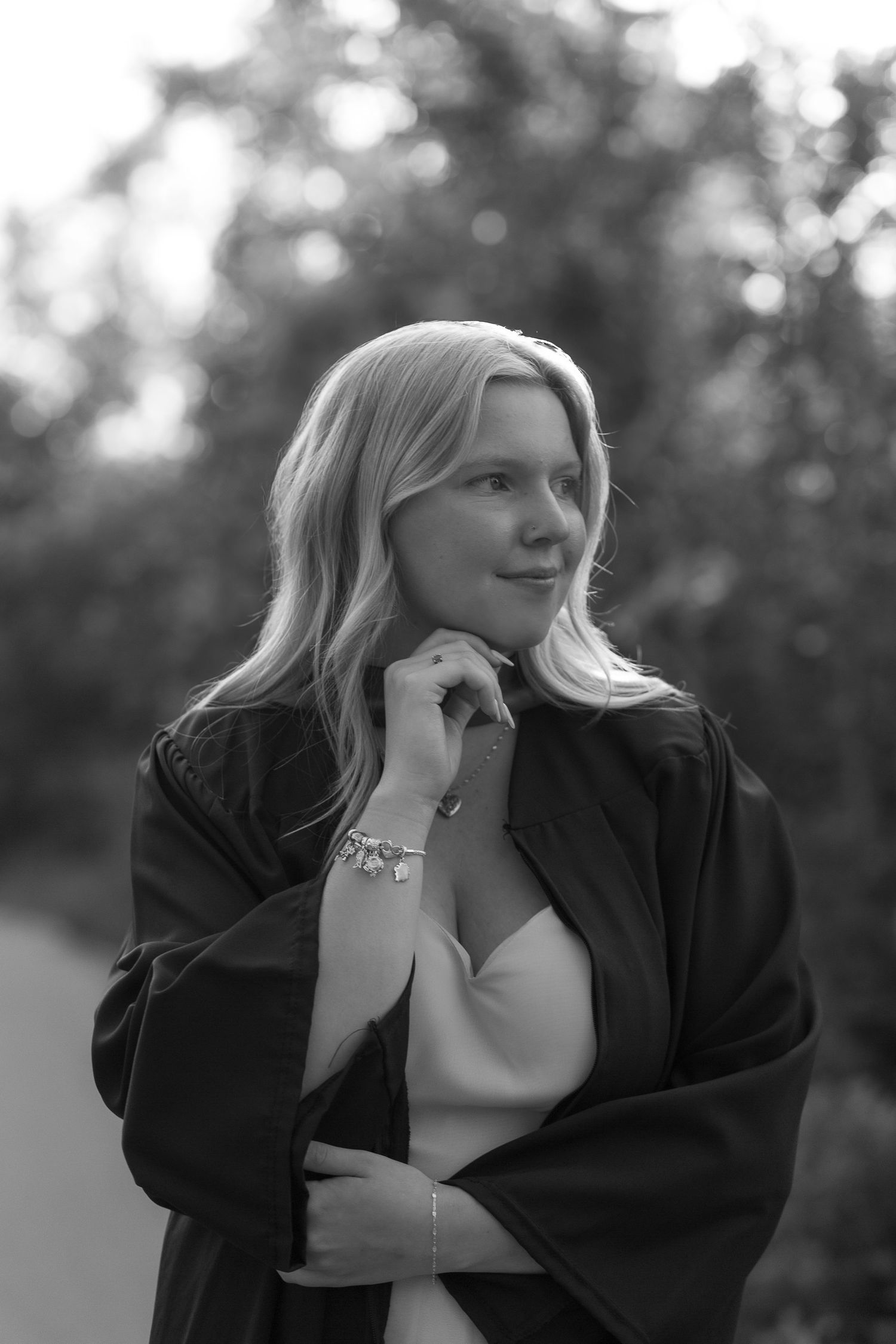 Black and white portrait of a blonde woman in a dark coat and bracelet, posing outdoors with blurred trees behind her.